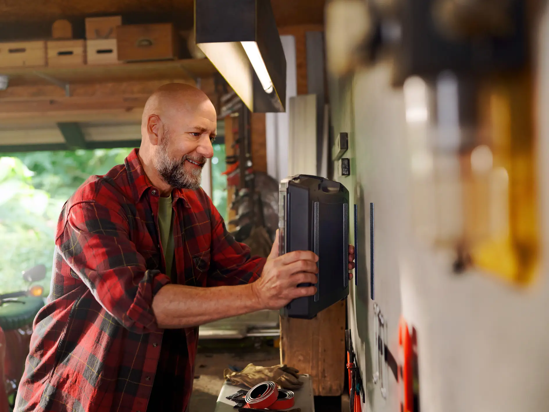 A bald man with a beard, wearing a red plaid shirt, is placing a toolbox on a shelf in his workshop. He smiles as he organizes tools around him. The workshop is brightly lit with various tools and equipment visible in the background, along with tesa tape prominently displayed among them. (This text has been generated by AI)