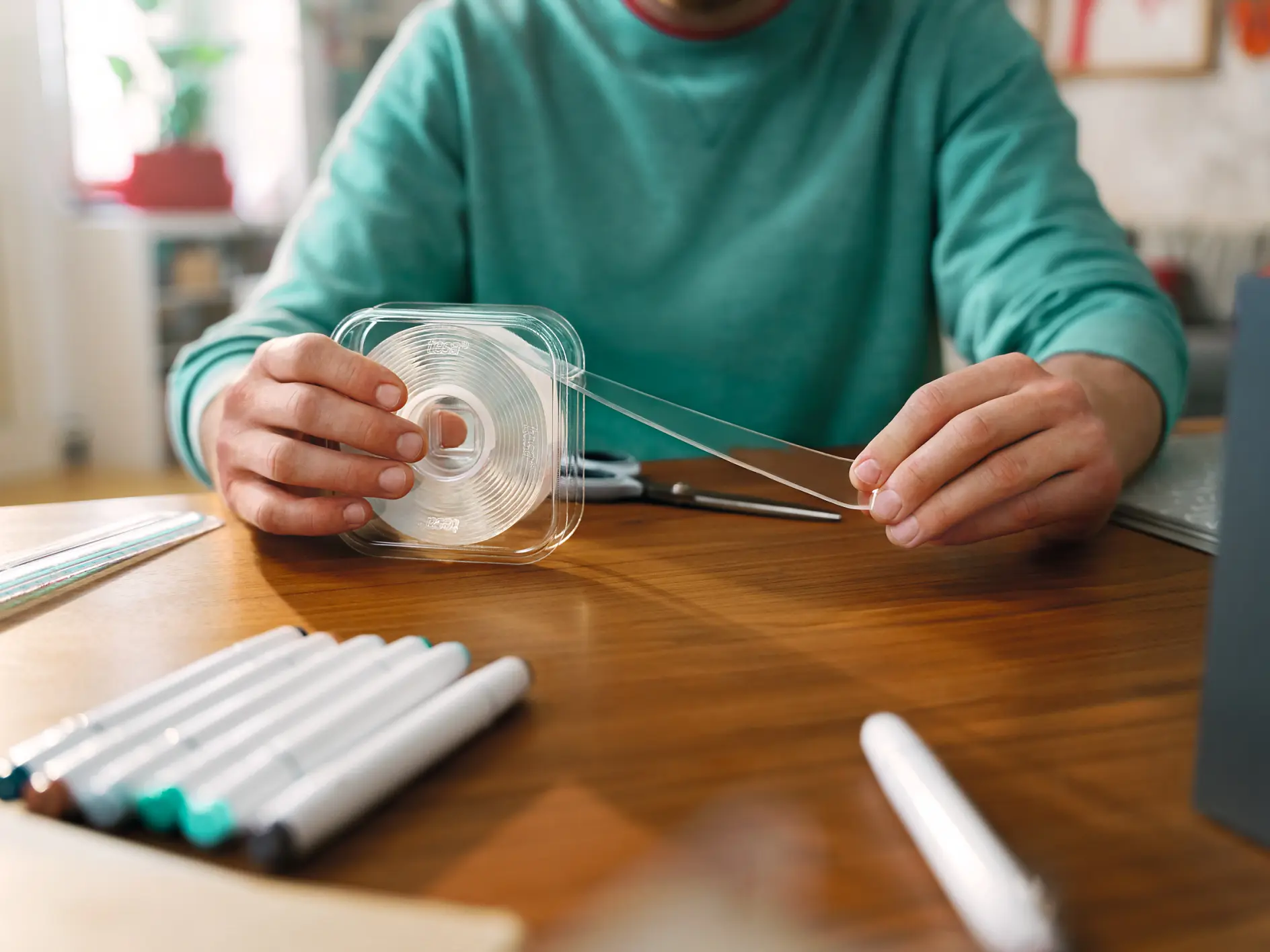 A person wearing a green shirt is seated at a wooden table, handling a clear tesa tape dispenser. Several markers are scattered on the table, and a notebook is partially visible. Scissors are also present near the persons hands. (This text has been generated by AI)