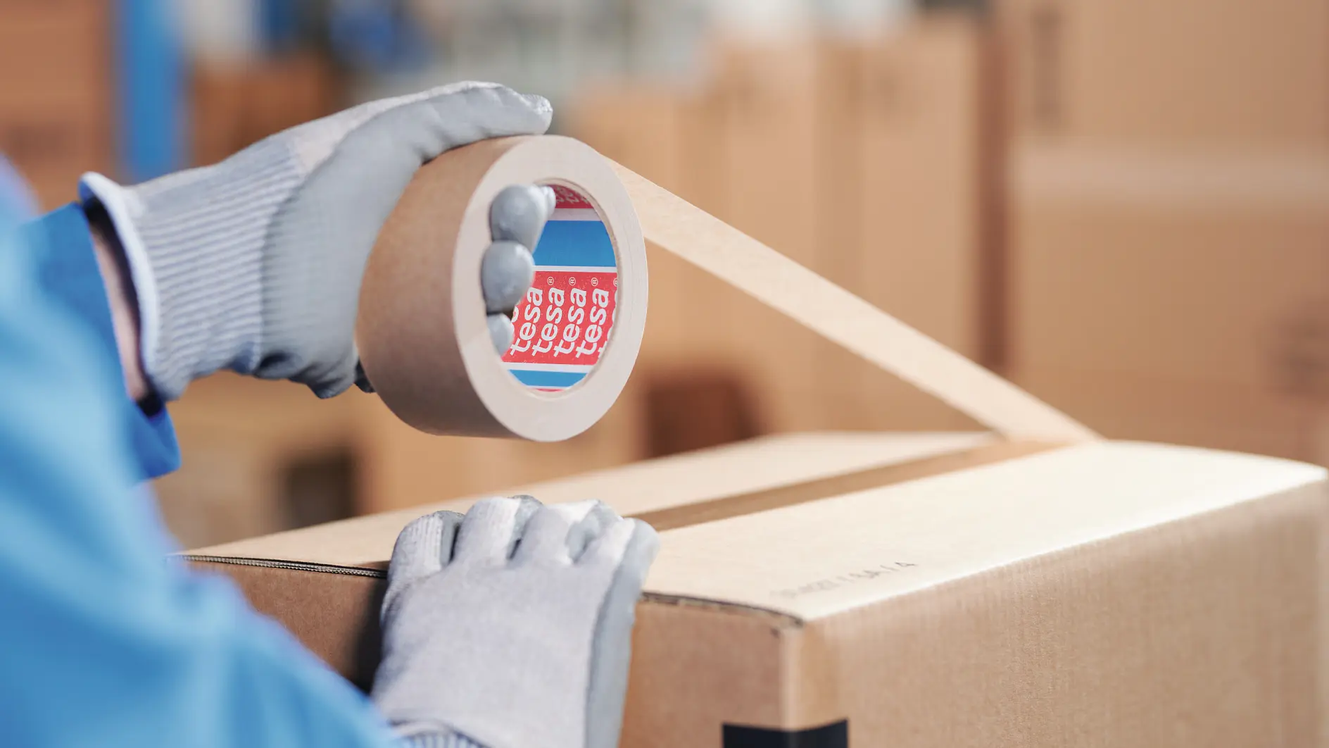 person wearing gloves sealing a cardboard box with clear tesa tape in a warehouse setting (This text has been generated by AI)