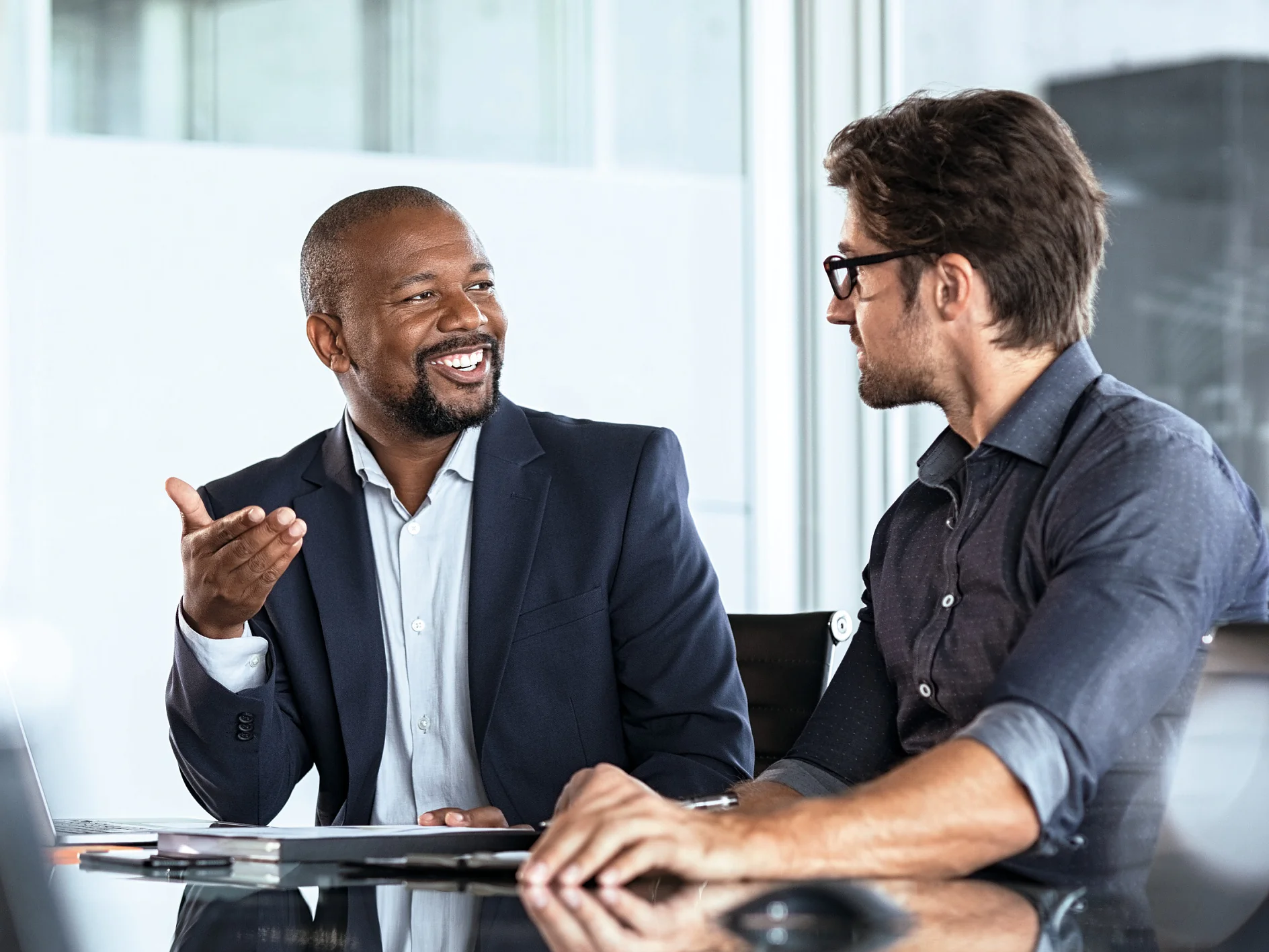 Two men in business attire engaged in discussion at an office desk with laptop and documents, indoor professional setting. (This text has been generated by AI)