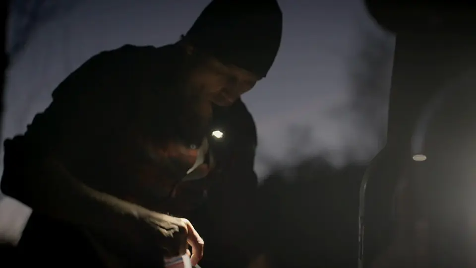 Άτομο που εργάζεται σε εξωτερικό χώρο το σούρουπο, φωτισμένο από ένα μικρό μπροστινό φως, ενώ χειρίζεται ένα εργαλείο ή αντικείμενο δίπλα σε μια έντονη λάμπα.