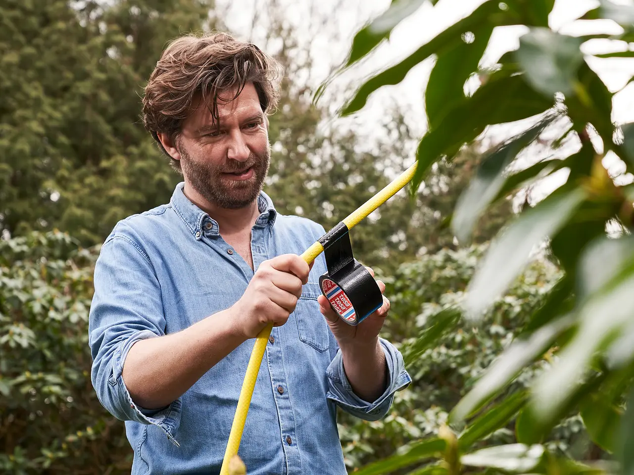 Ένας γενειοφόρος άνδρας με μπλε πουκάμισο στέκεται έξω, κρατώντας ένα κίτρινο λάστιχο και ένα ρολό ταινίας tesa. Φαίνεται να επισκευάζει ή να ρυθμίζει τον εύκαμπτο σωλήνα. Πράσινο φύλλωμα και δέντρα τον περιβάλλουν στο βάθος. (This text has been generated by AI)