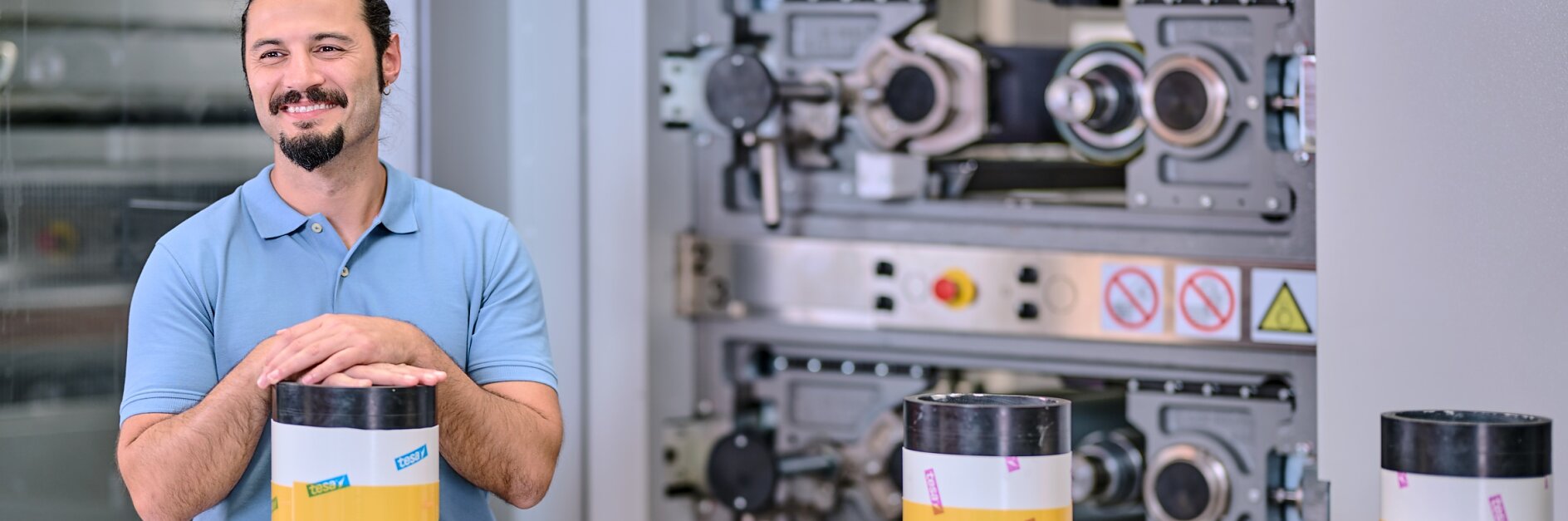 Ένας άνδρας με γένια και αλογοουρά, φορώντας μια μπλε μπλούζα πόλο, στέκεται χαμογελαστός με τα χέρια του πάνω σε ένα μεγάλο κυλινδρικό αντικείμενο τυλιγμένο με κίτρινη ταινία tesa. Βιομηχανικά μηχανήματα διακρίνονται στο φόντο.