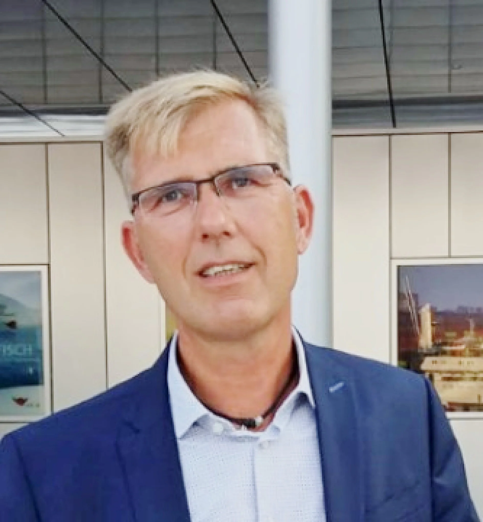 A man with short blond hair and glasses is wearing a blue suit jacket over a white shirt. He is standing indoors in front of a wall with framed pictures, where several items are affixed using tesa tape. (This text has been generated by AI)