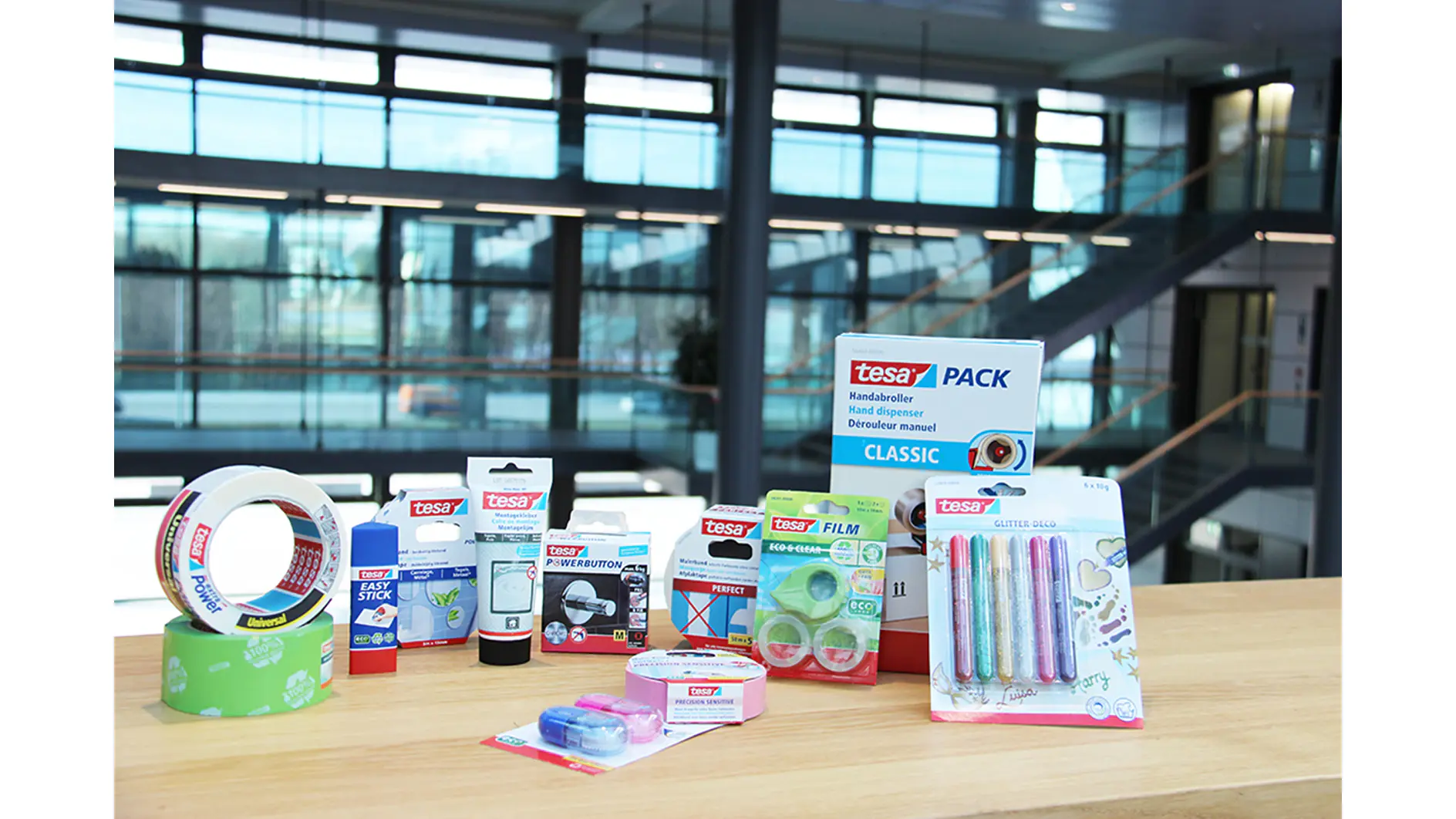 Various Tesa brand stationery items, including tesa tape, glue sticks, correction tape, and pens, are displayed on a wooden surface. A modern office with large windows and stairs is visible in the background. (This text has been generated by AI)