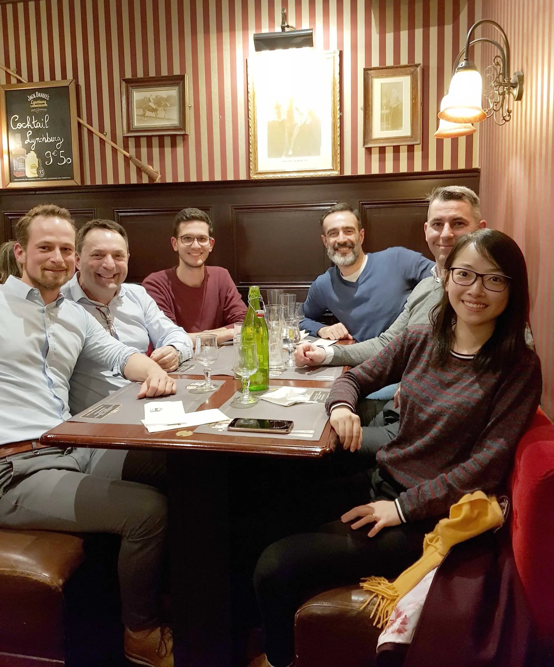 Six people are sitting around a rectangular table in a cozy restaurant with red and white striped walls. They are smiling at the camera. A green bottle and glasses are on the table. Framed pictures and a chalkboard are on the wall behind them, secured carefully with tesa tape.