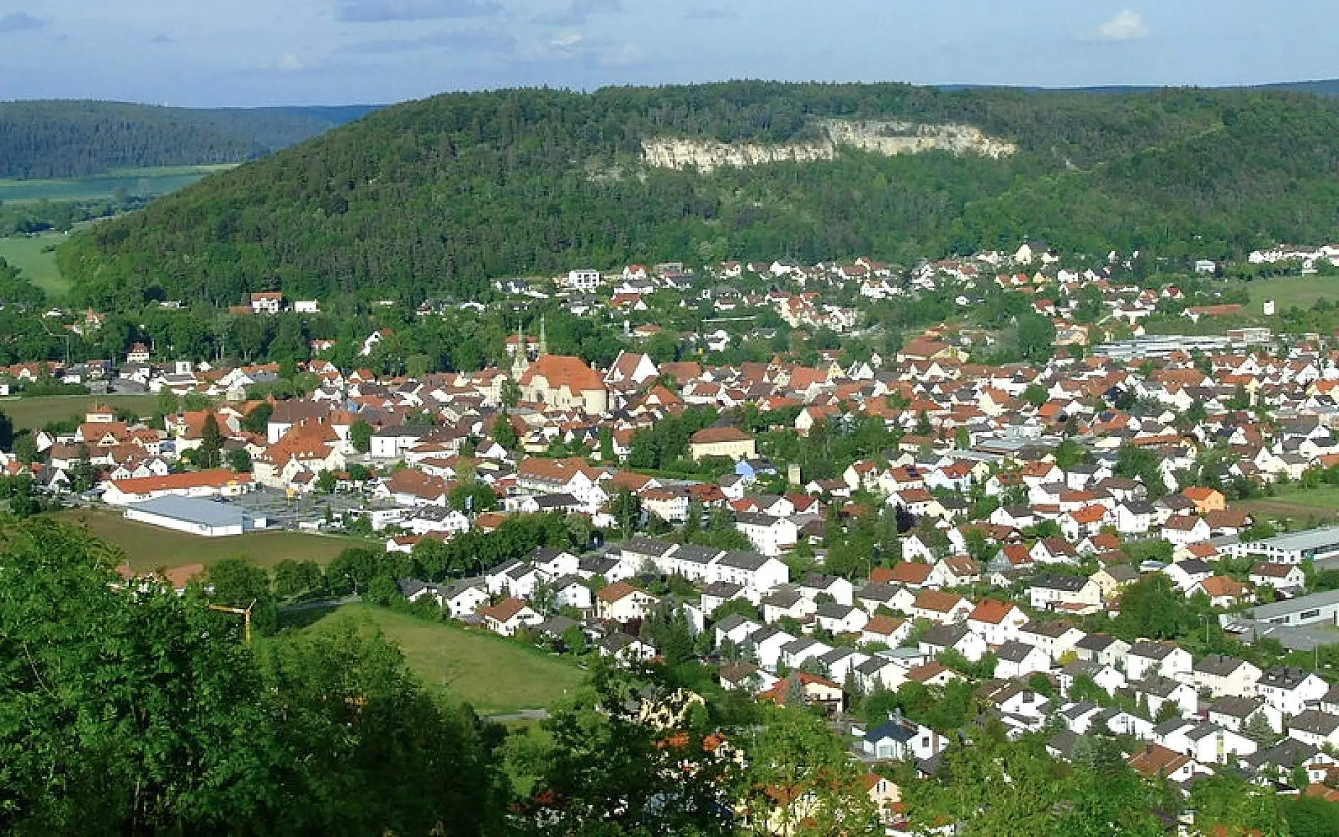 Aerial view of a small town with red-roofed buildings surrounded by lush green hills and forested areas. The town appears to be nestled in a valley, with several clustered residential and community structures visible.