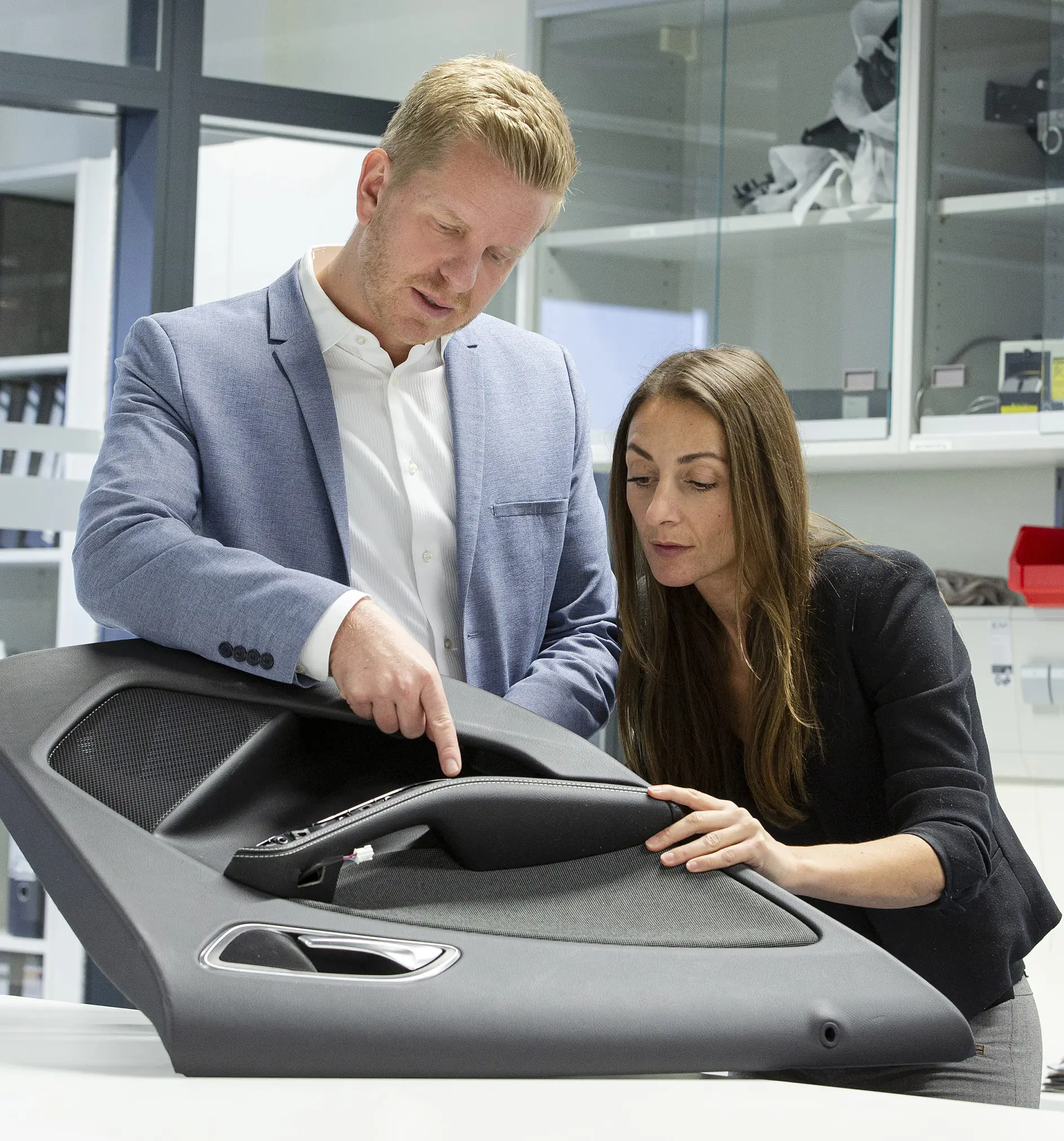 A man and woman examine an interior car door panel in an office setting. The man points at the panel while the woman touches it. Shelves with various items, including rolls of tesa tape, are visible in the background. Both are dressed in business attire.
