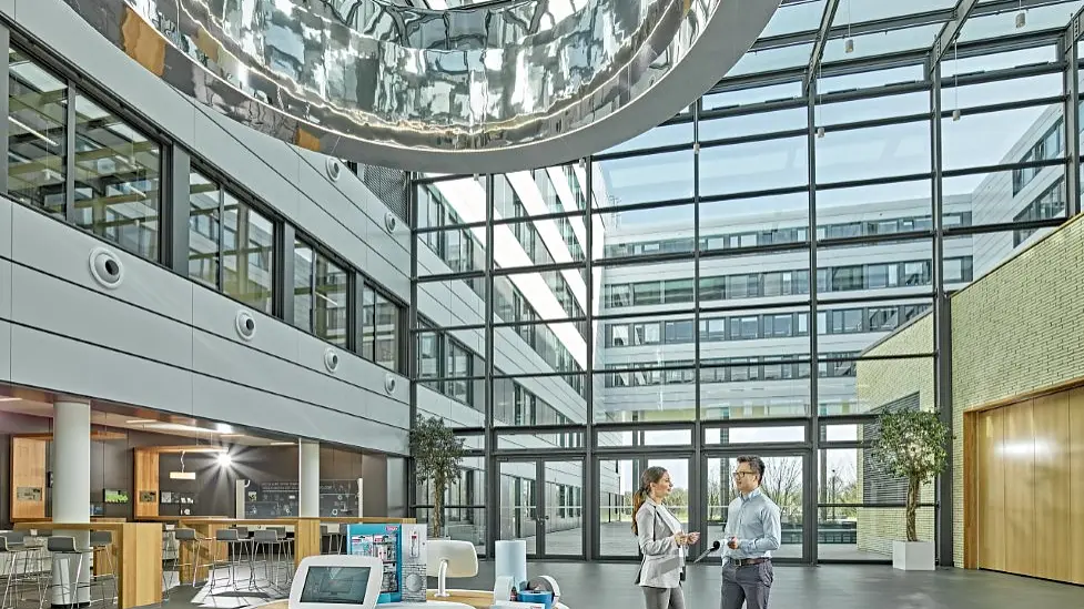 A modern, spacious lobby with a glass ceiling and a large circular metal sculpture. Two people stand talking near a round display table with electronic devices secured by tesa tape. Potted trees and a seating area are visible. Natural light fills the space. (This text has been generated by AI)