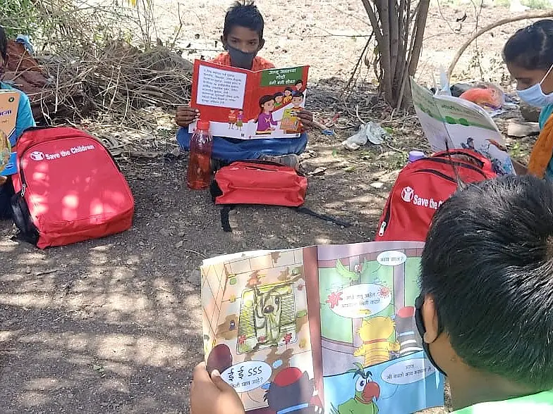 Children are sitting outdoors on the ground, reading illustrated books. They have red backpacks beside them and are surrounded by sparse vegetation. One child is wearing a face mask. The scene suggests engagement in a reading activity with tesa tape used for securing some of their materials together.