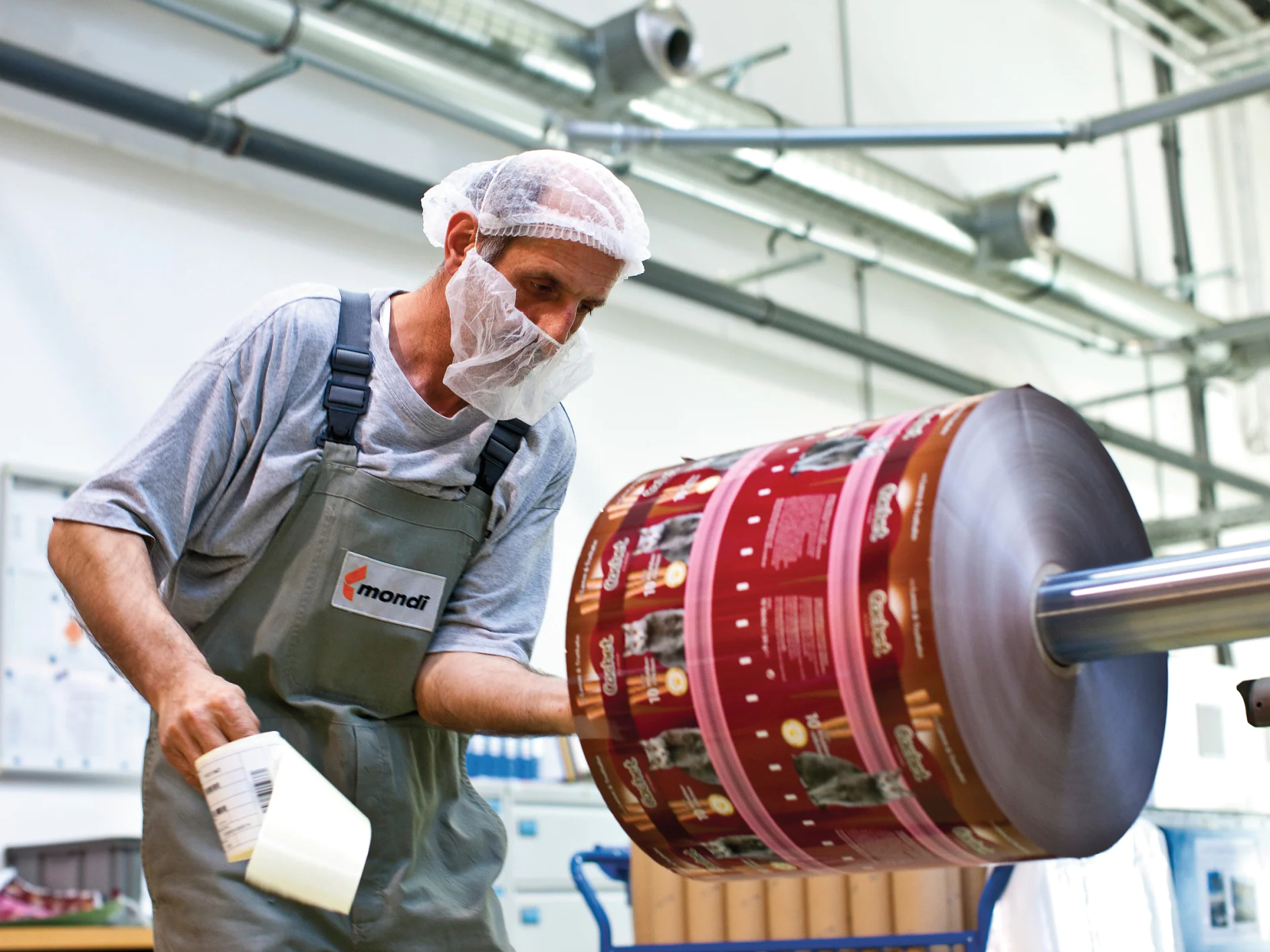 A worker wearing a hairnet and face mask inspects a large roll of printed packaging material in a factory setting. He holds a tool and is dressed in a gray shirt and green overalls. Machinery, tesa tape, and industrial elements are visible in the background. (This text has been generated by AI)