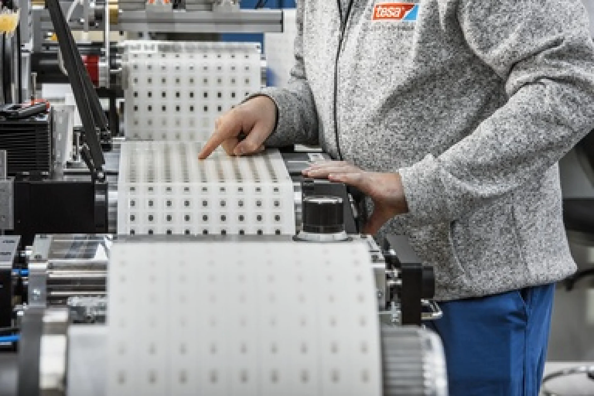 A person wearing a gray sweater operates a machine with large rolls of tesa tape, featuring square cutouts. The workspace appears industrial, with a focus on precision handling of the material. (This text has been generated by AI)