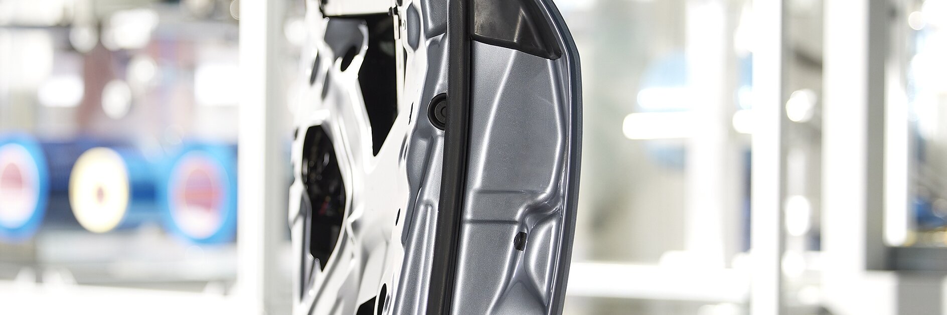 Side view of a metal car door frame in a manufacturing setting. The frame is silver with visible cutouts and structural details, secured with tesa tape. The background is blurred, featuring industrial equipment and machinery.