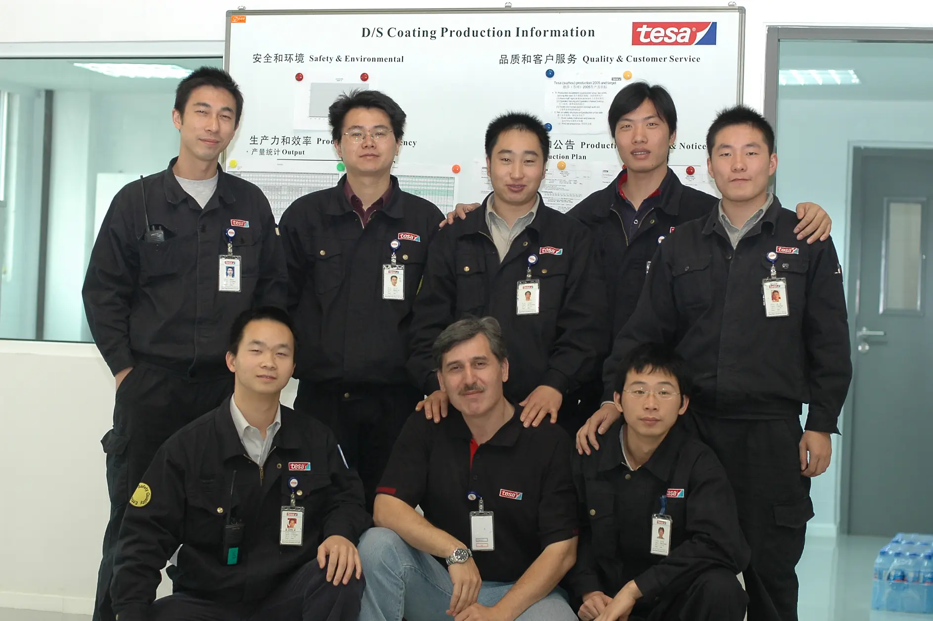 A group of nine men in matching black uniforms pose for a photo in front of a poster labeled D/S Coating Production Information. They are standing and kneeling in a well-lit room with white walls and a gray door, all proudly showcasing products from tesa tape.