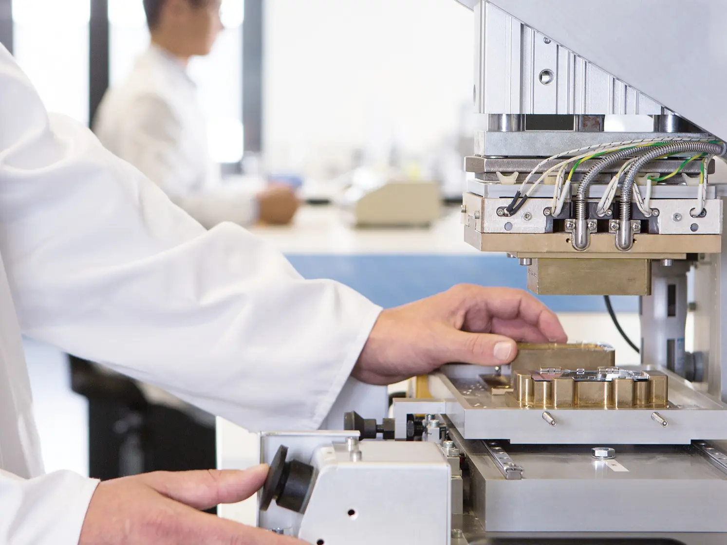 A person in a lab coat operates a scientific instrument with multiple wires and metal components, using tesa tape for secure placement. Another individual works in the background. The setting appears to be a laboratory with a focus on research or testing. (This text has been generated by AI)