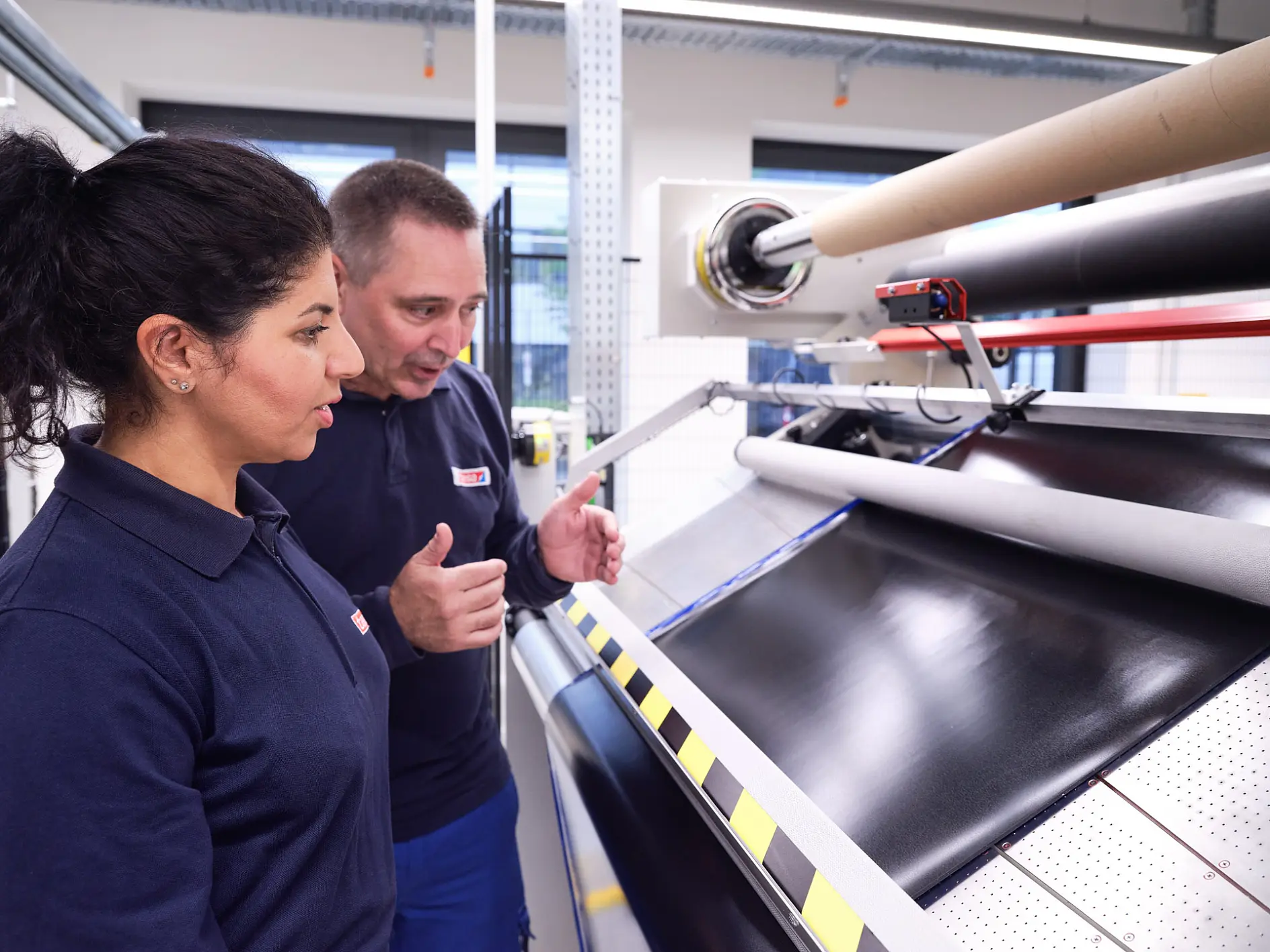 Two workers in a factory setting are observing a large sheet of black material being processed on a machine. The man explains something to the woman, demonstrating how tesa tape is used in the process. Both are wearing dark blue uniforms with red and white patches. The room is illuminated with industrial lighting. (This text has been generated by AI)