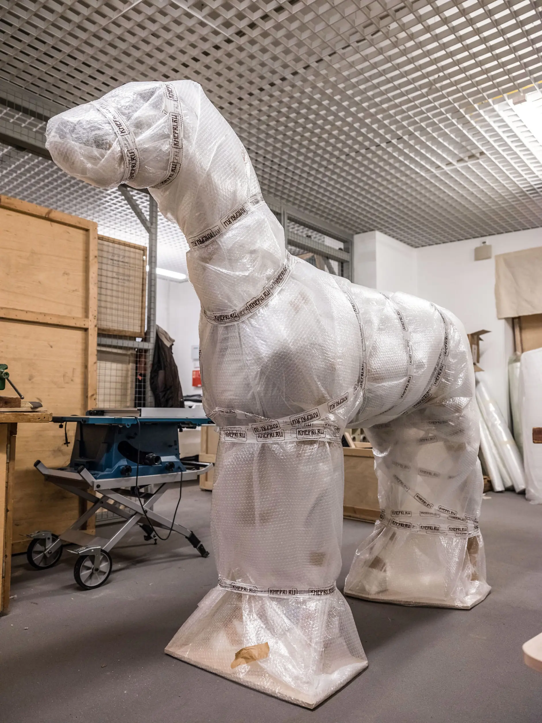 A large sculpture wrapped in bubble wrap and tesa tape resembles a horse with an elongated neck. The scene is set in an indoor workspace with wooden crates and various tools in the background.