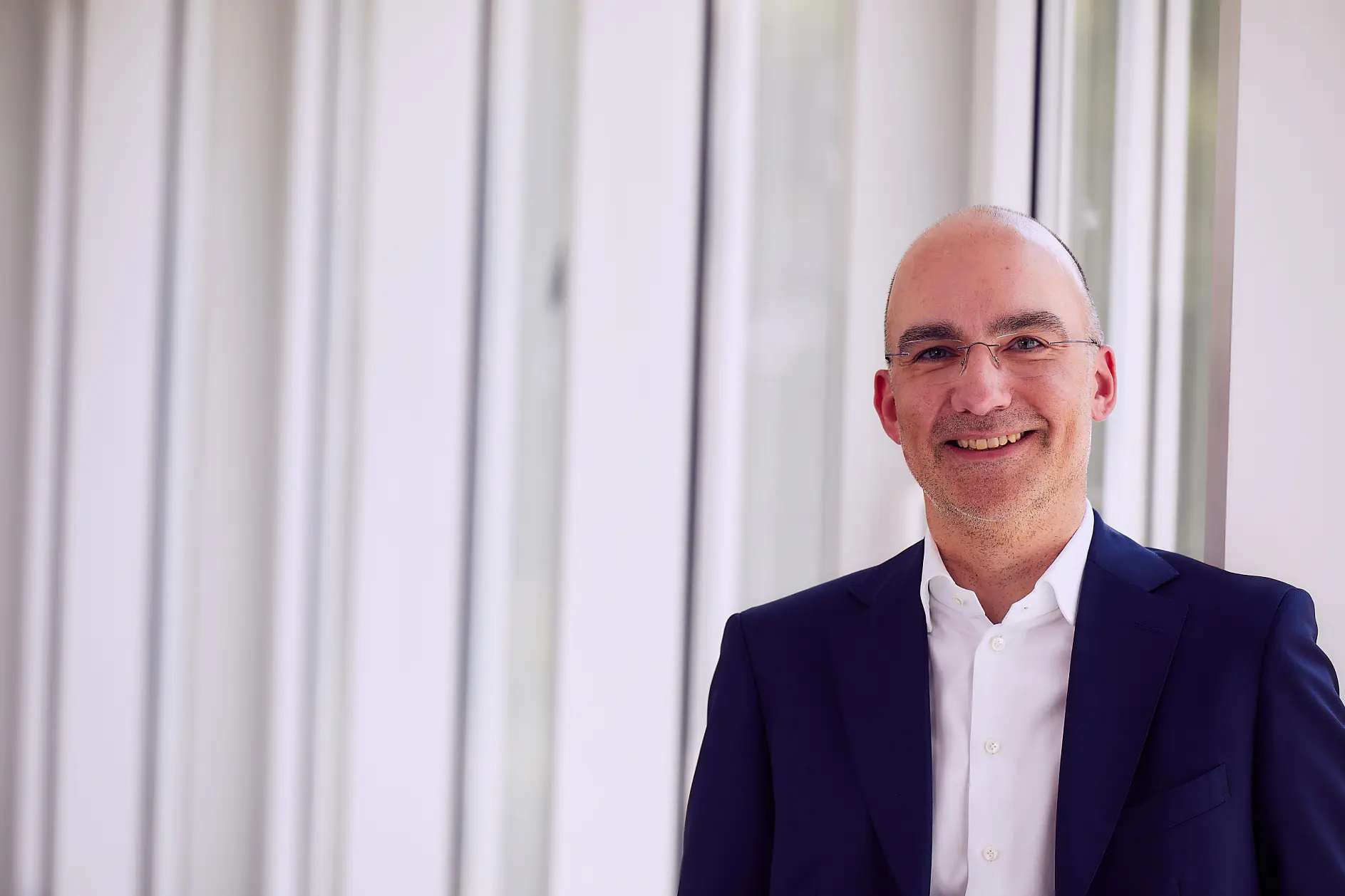 A bald man with glasses is smiling, wearing a white shirt and a dark suit jacket. He stands near a light-colored wall with vertical panels, where pieces of tesa tape hold various items in place.