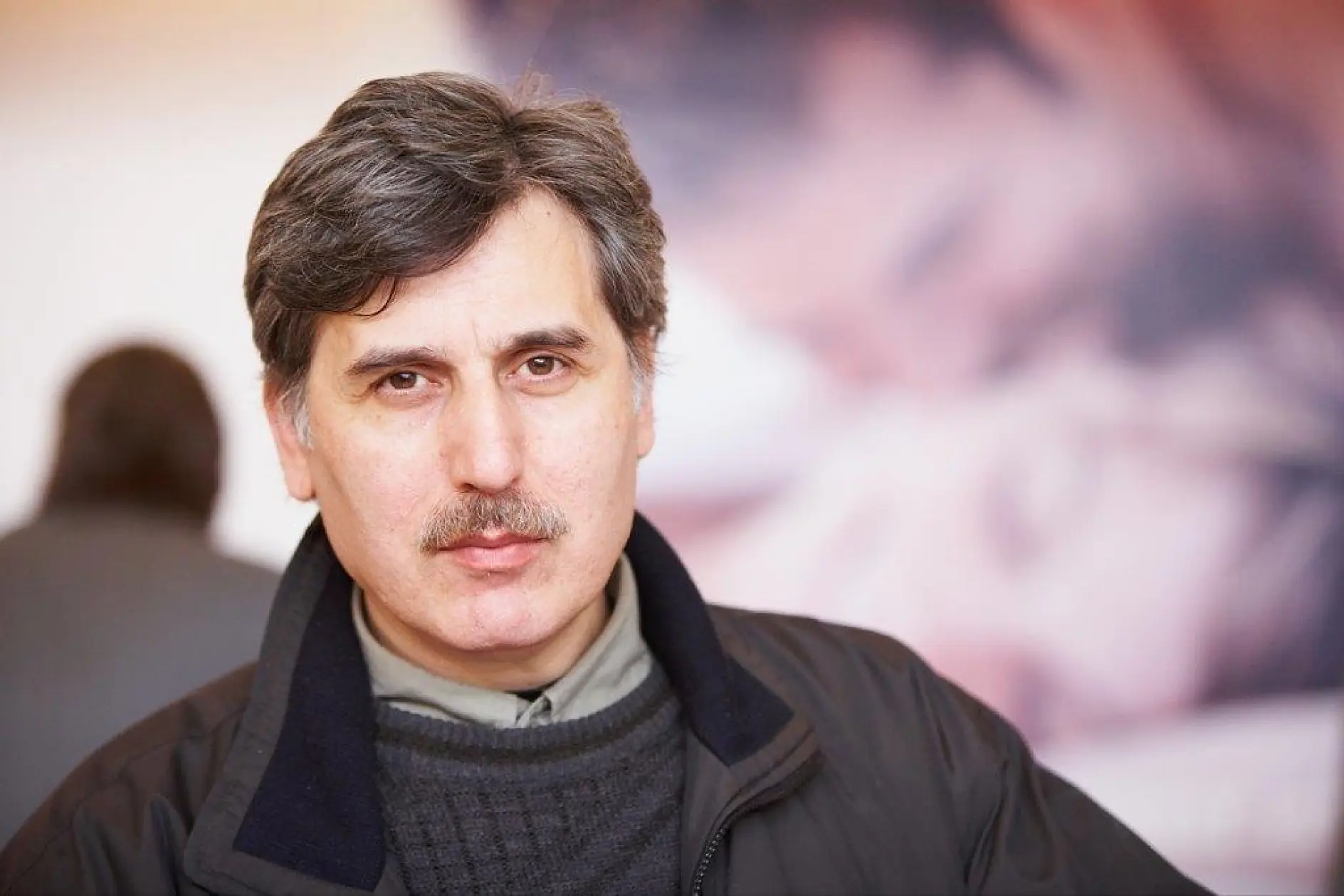 A man with gray hair and a mustache is looking at the camera. He is wearing a black jacket over a gray shirt. The background is blurred, with what appears to be another person in the distance, possibly using tesa tape.