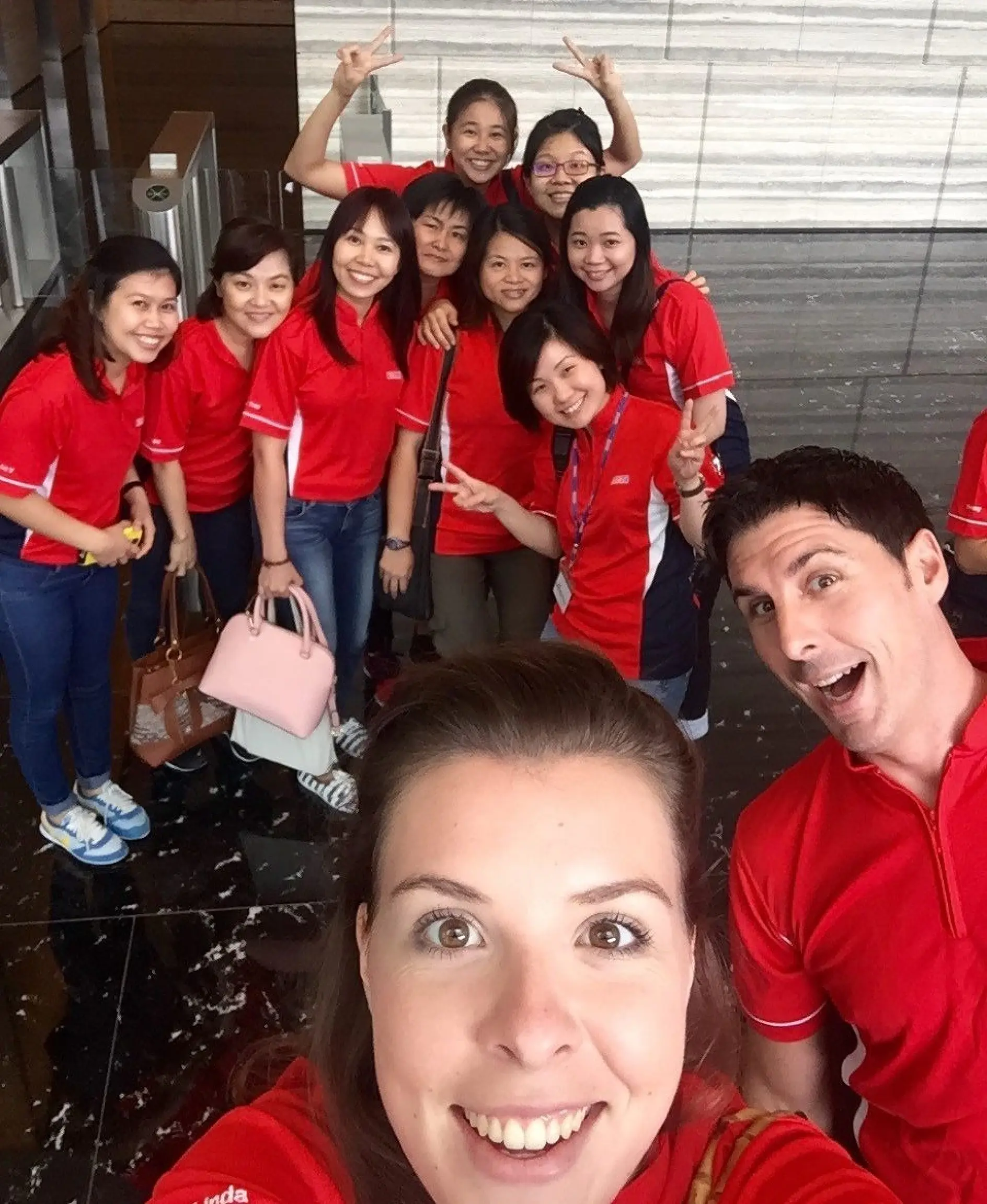 A group of people wearing red shirts are posing indoors. A woman and a man in the foreground are smiling at the camera. The rest of the group is behind them, some making peace signs. The floor is shiny and reflective, secured with tesa tape for added durability.