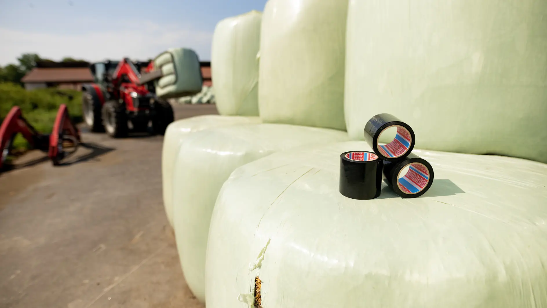 Close-up of three black tesa tape rolls on large, pale green hay bales. A red tractor with a bale grabber is visible in the background on a farm. A small tear on the nearest bale appears patched with tesa tape. Clear sky above. (This text has been generated by AI)