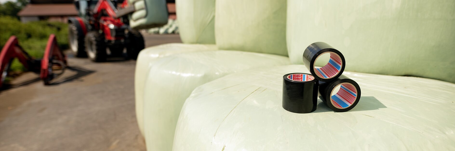 Close-up of three black tesa tape rolls on large, pale green hay bales. A red tractor with a bale grabber is visible in the background on a farm. A small tear on the nearest bale appears patched with tesa tape. Clear sky above.