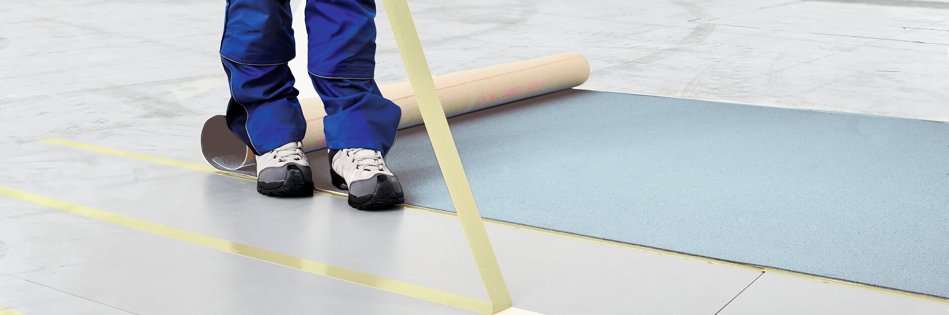 A person wearing blue pants and sneakers is applying yellow tesa tape to a light gray floor in a large indoor space. A roll of tesa tape is visible on the floor, partially unrolled onto the surface.