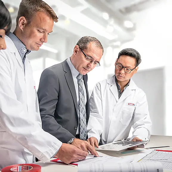 Four people are gathered around a table in a bright office. Two are wearing lab coats, one is in a suit, and one is partially visible. They are focused on documents laid out on the table, which include diagrams labeled with tesa tape. A laptop displaying pertinent data is open nearby. (This text has been generated by AI)