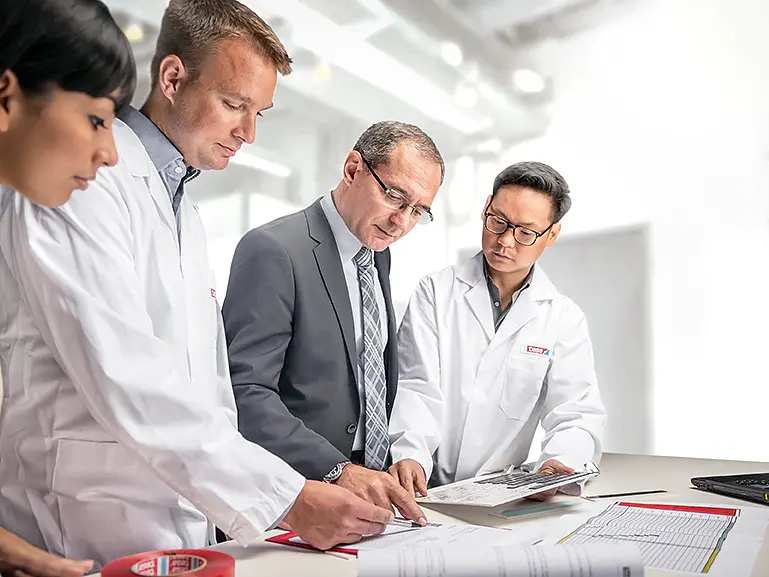 Four people are gathered around a table in a bright office. Two are wearing lab coats, one is in a suit, and one is partially visible. They are focused on documents laid out on the table, which include diagrams labeled with tesa tape. A laptop displaying pertinent data is open nearby. (This text has been generated by AI)