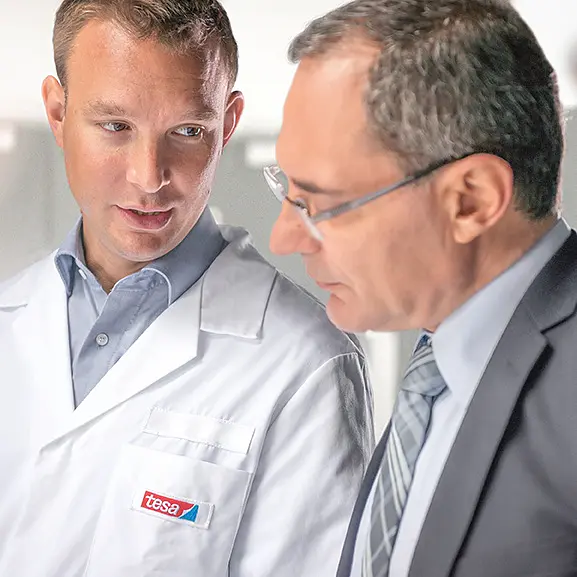 A man in a white lab coat with a tesa tape label talks to another man in a suit and glasses. They appear to be in a discussion while looking at something out of the frame. The background is bright and slightly blurred. (This text has been generated by AI)