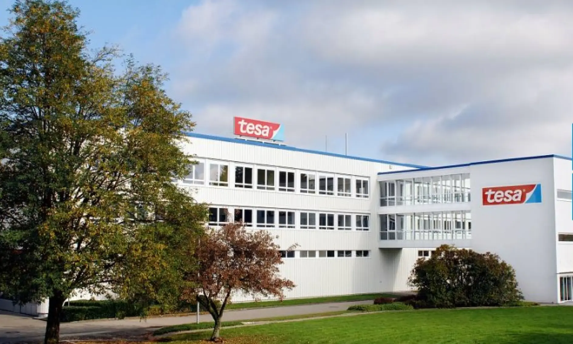 A large, white, multi-story building with several windows, displaying the red and white tesa tape logo on the roof and side. The building is surrounded by trees and grass, with a partly cloudy sky above.