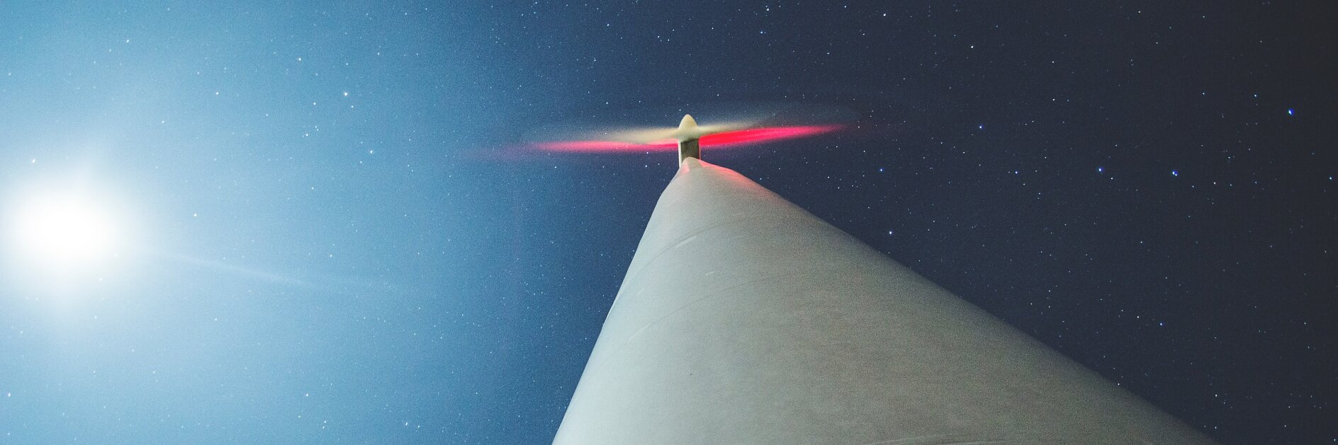 Looking up at a wind turbine against a starry night sky with the full moon visible. The turbines blades have a slight red glow, and its base is marked with V 24070.