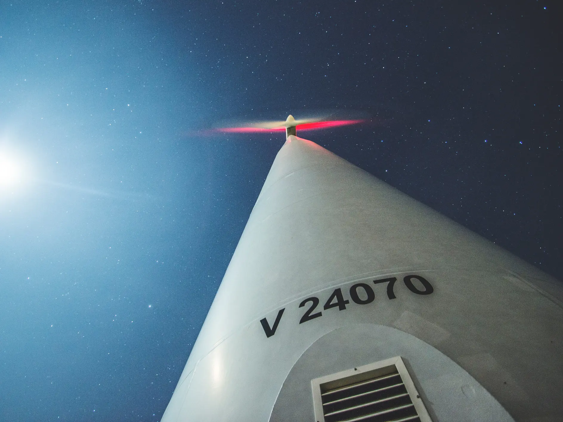 Looking up at a wind turbine against a starry night sky with the full moon visible. The turbines blades have a slight red glow, and its base is marked with V 24070. (This text has been generated by AI)