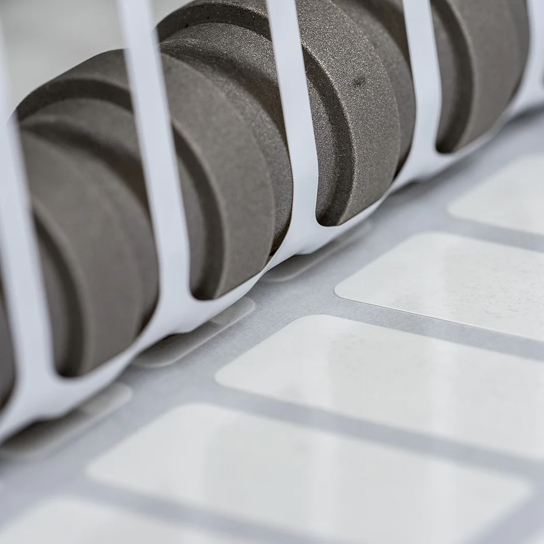 Close-up of a set of black gears positioned next to a row of white labels running along a conveyor system, partially enclosed by white vertical bars. The scene suggests an industrial setting, likely related to printing or labeling using tesa tape. (This text has been generated by AI)