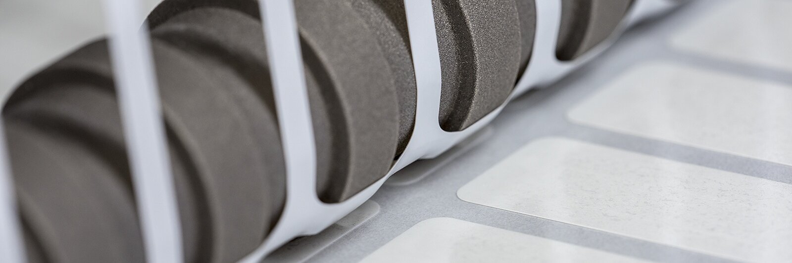 Close-up of a set of black gears positioned next to a row of white labels running along a conveyor system, partially enclosed by white vertical bars. The scene suggests an industrial setting, likely related to printing or labeling using tesa tape. (This text has been generated by AI)