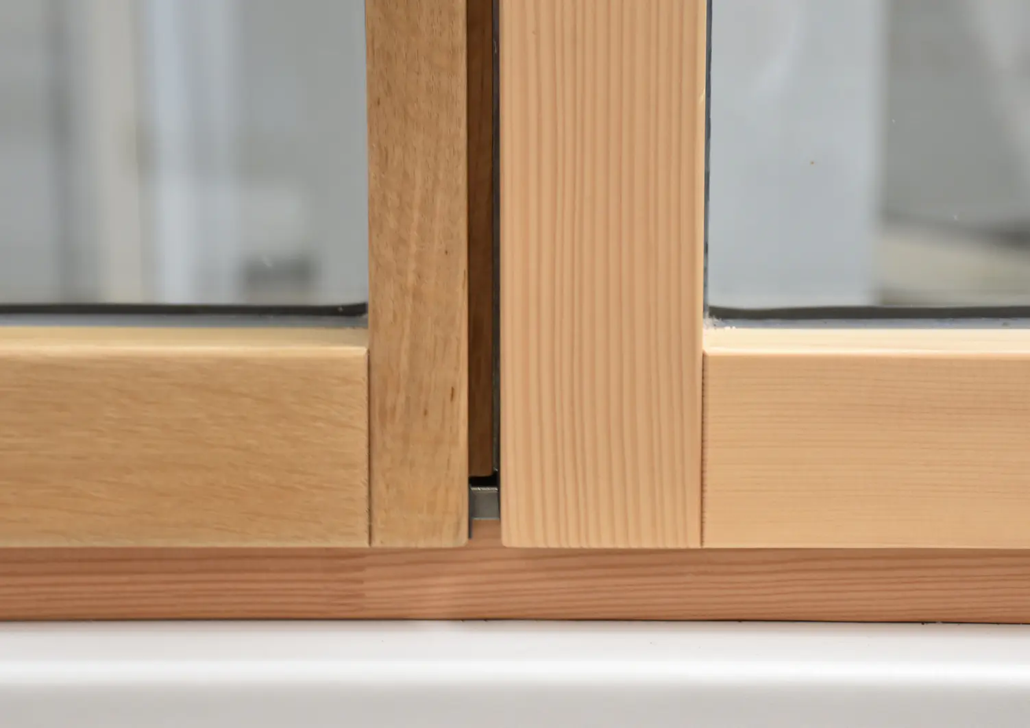 Close-up of two wooden window frames with clear glass panes. The frames have a natural wood finish and meet in the middle, secured with tesa tape. The background is out of focus, highlighting the wood grain texture and the joint where the frames converge. (This text has been generated by AI)