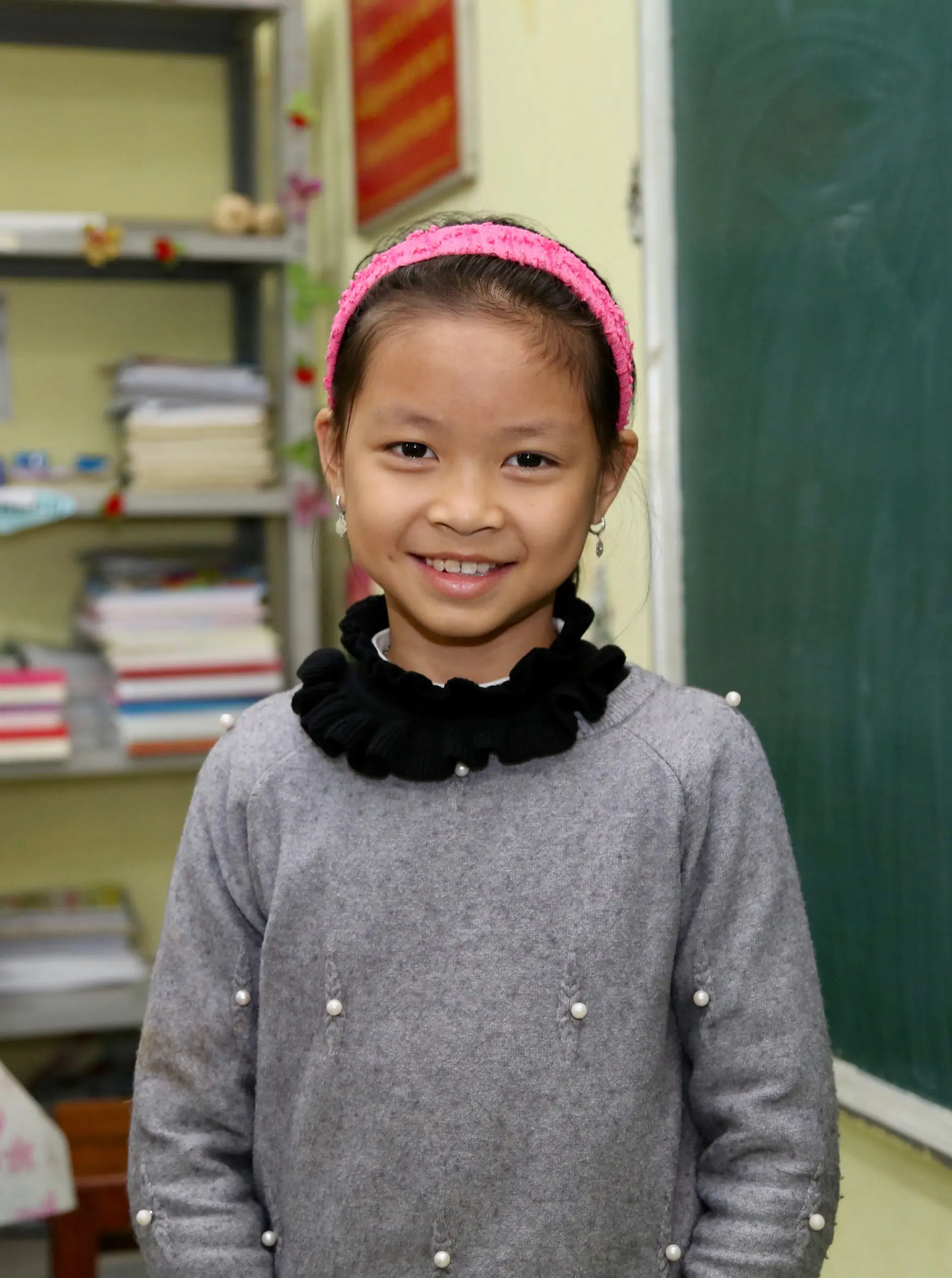 A young girl wearing a gray sweater with pearl details and a pink headband is smiling at the camera. She stands in a classroom in front of a chalkboard, with stacks of books on a shelf in the background, ready for her next project using tesa tape.