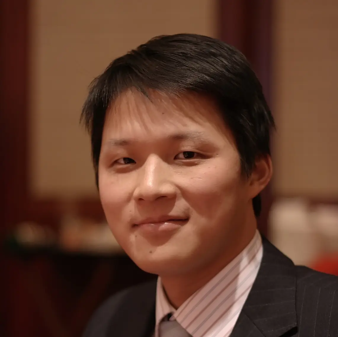 A person wearing a dark suit, light striped shirt, and a patterned tie is sitting indoors. The background is softly blurred, highlighting the individual as the main focus. The lighting is warm, giving the scene a professional and formal atmosphere.