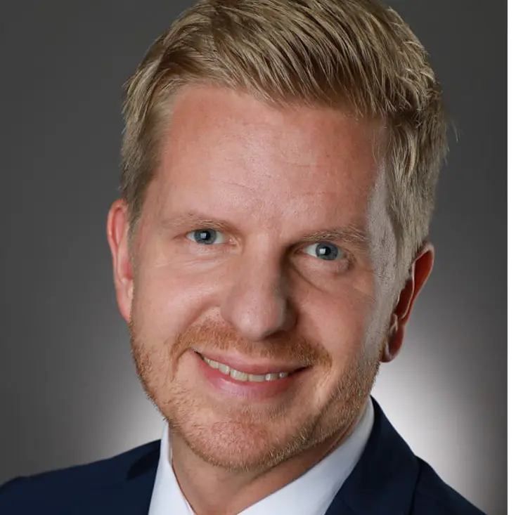 A person with short blond hair and a beard is wearing a navy blue suit, white shirt, and red tie. Smiling at the camera, the background is gray with a subtle spotlight effect.