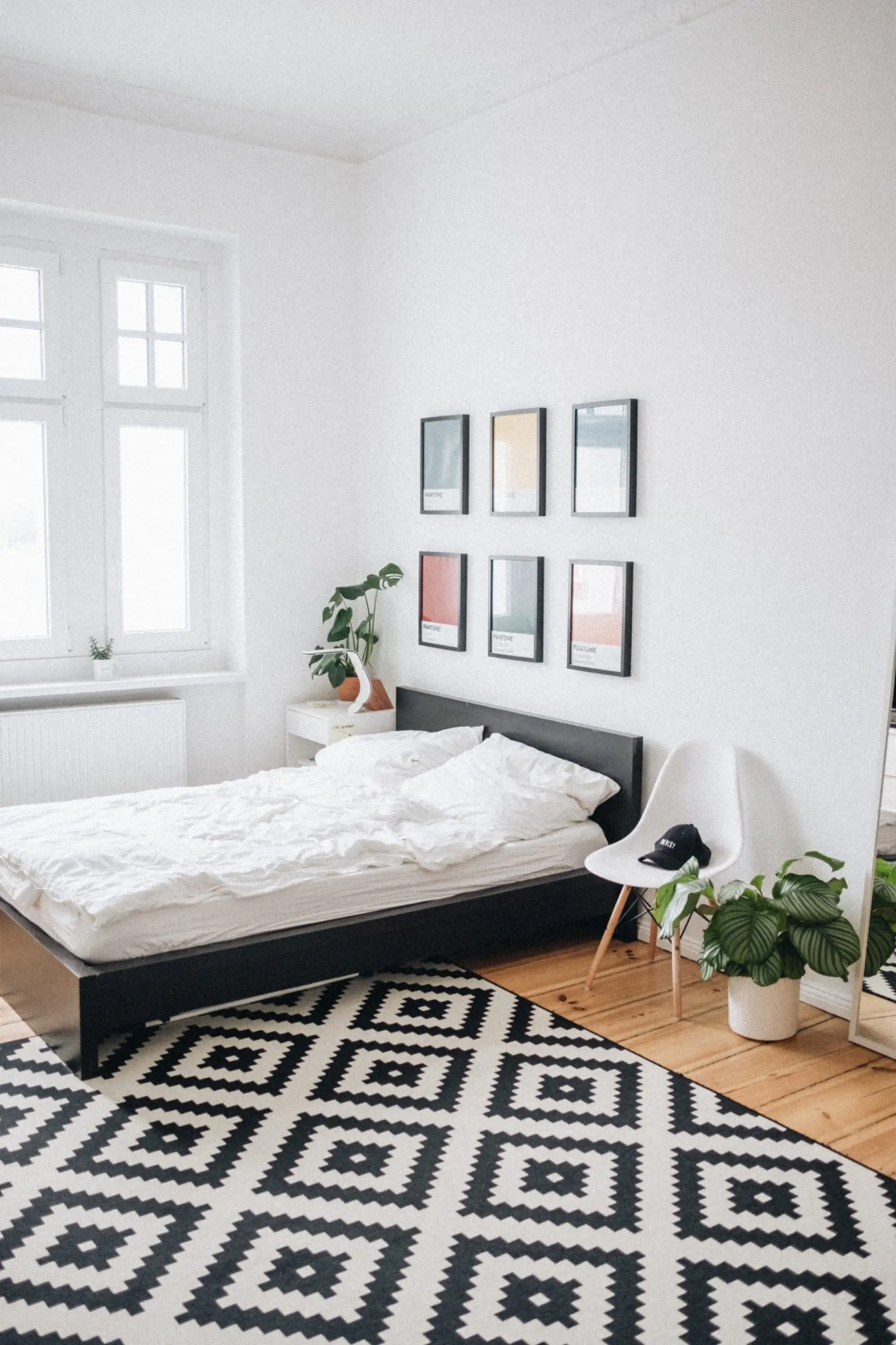 A bedroom features a black bed with white bedding and a geometric black and white rug. Above the bed, there are five framed pictures. A white chair holding a cap sits next to a potted plant. A window is on the left wall, while a mirror adorns the right wall.