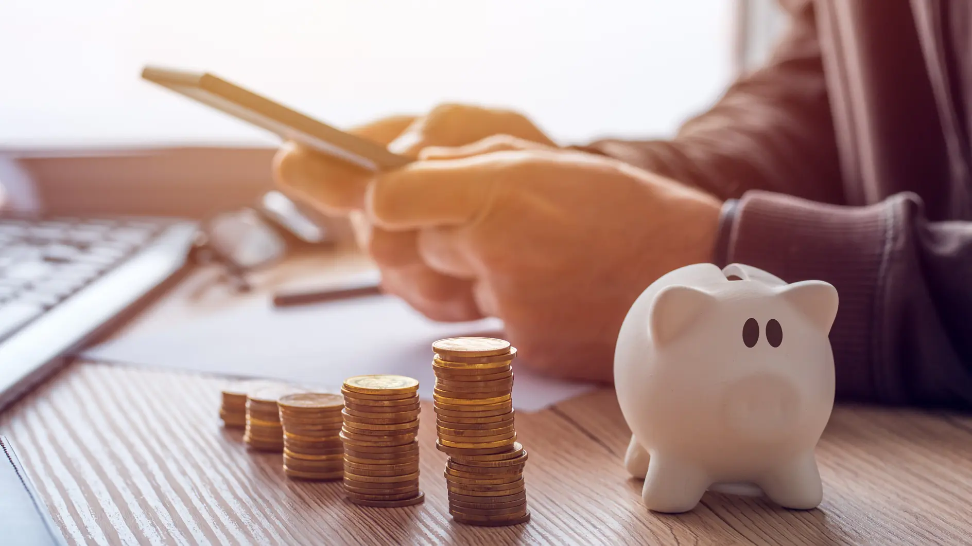 A person uses a smartphone at a desk with neatly stacked coins and a white piggy bank. The scene suggests financial planning or online banking, with a blurred keyboard and papers visible in the background.