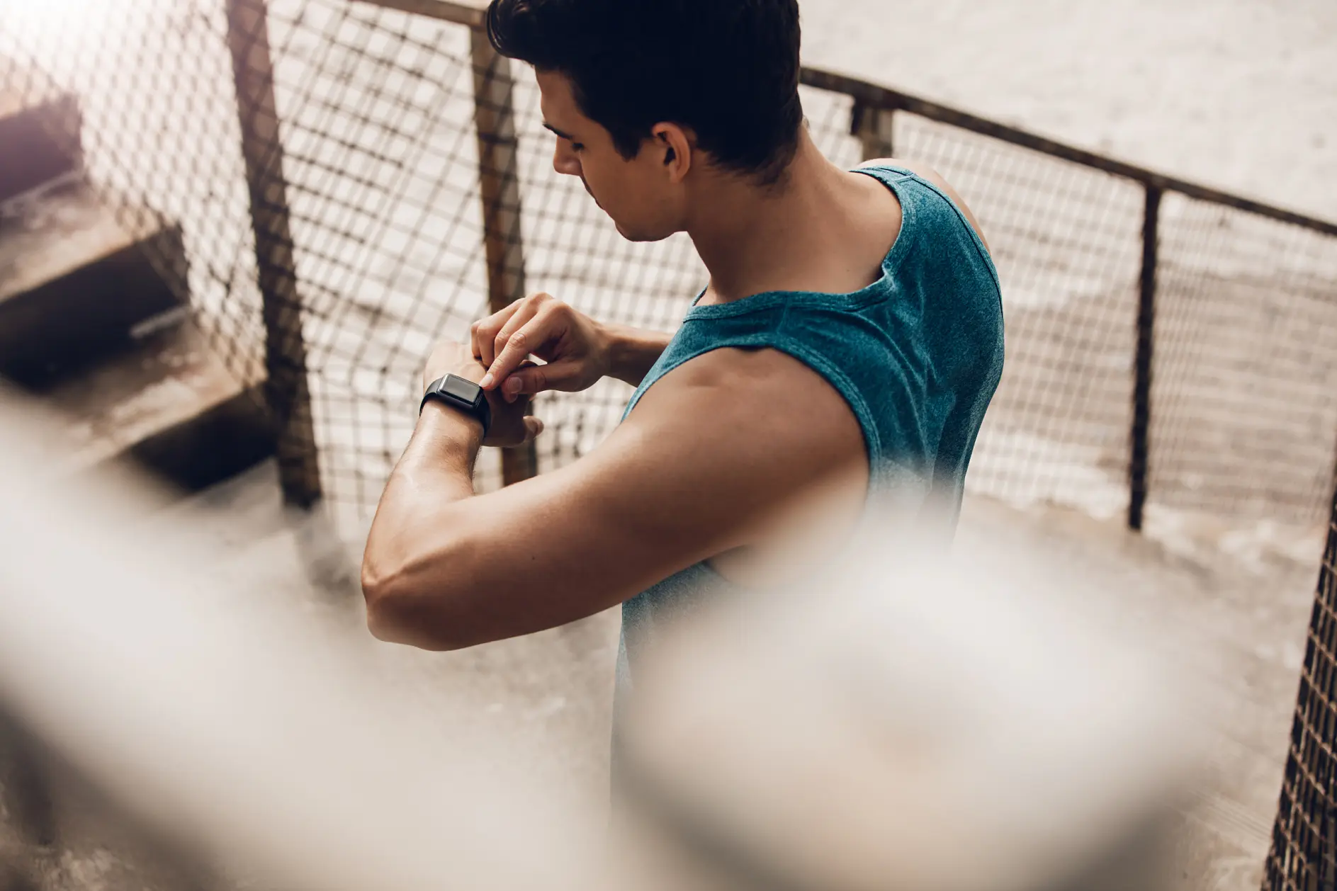 A man wearing a blue sleeveless shirt stands on an outdoor staircase, viewing a smartwatch on his left wrist. The scene is framed by a metal fence, and the ground appears concrete.