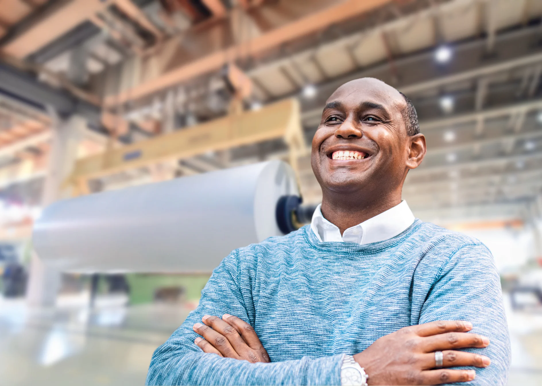 A person wearing a light blue sweater stands smiling with arms crossed in an industrial setting. The background features large rolls of paper and machinery, alongside various supplies of tesa tape. (This text has been generated by AI)