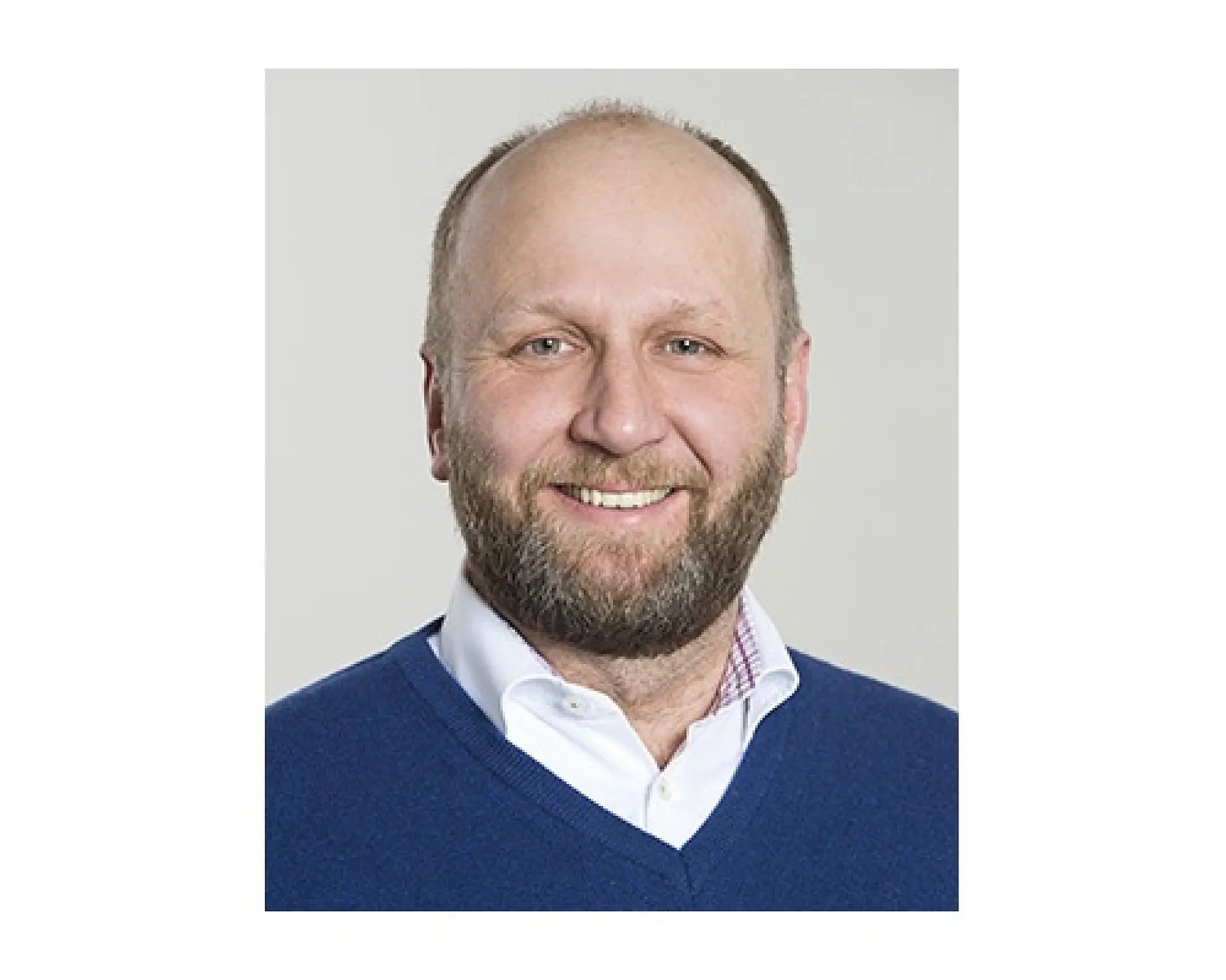 A person with a beard smiles for a portrait. They are wearing a white collared shirt and a blue sweater. The background is light gray, with no visible reference to tesa tape or any other products.