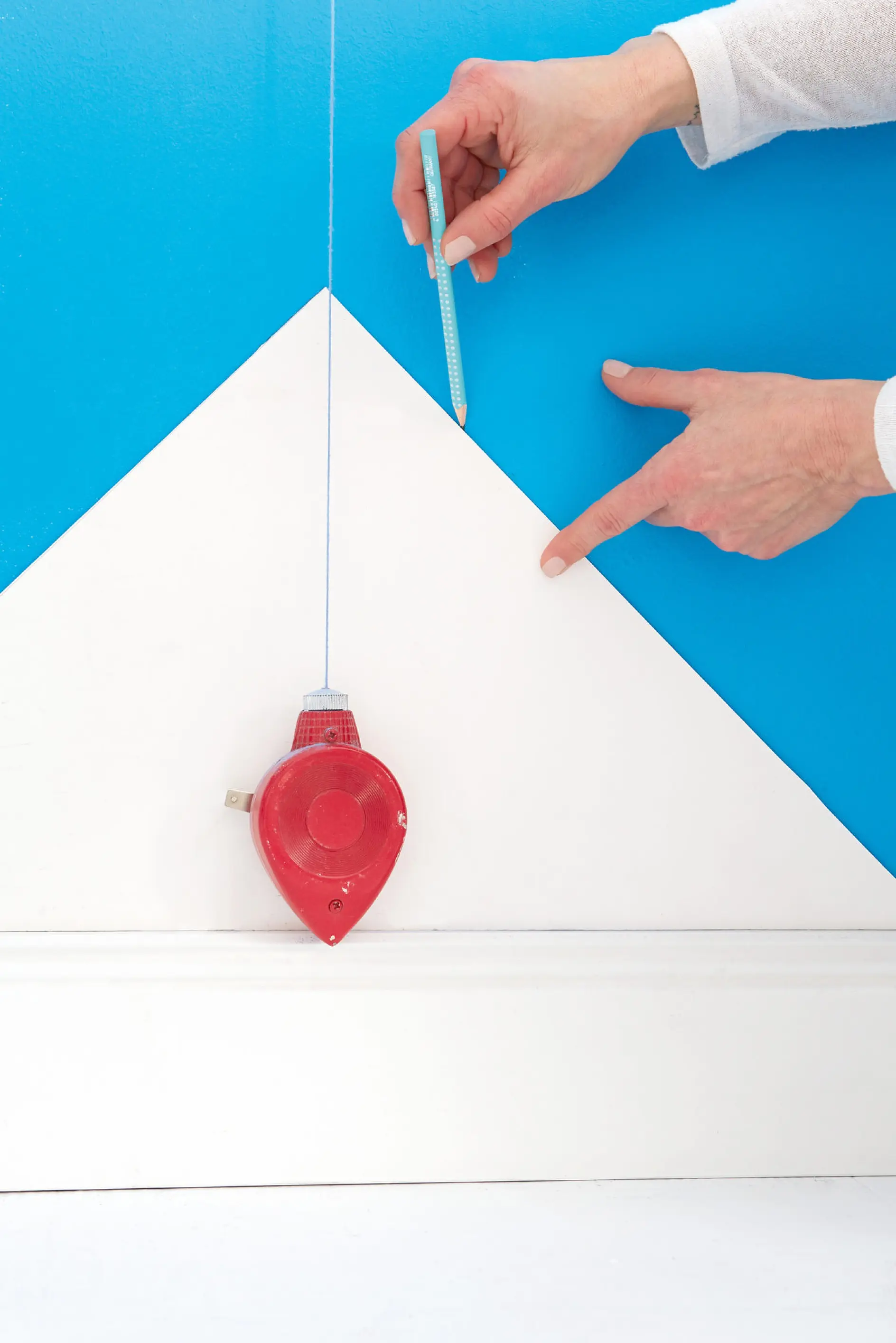 A person uses a red chalk line tool to mark a straight line on a blue and white wall. They hold a pencil and point at the intersection of the chalk line and the triangular white area, indicating a design or construction project.
