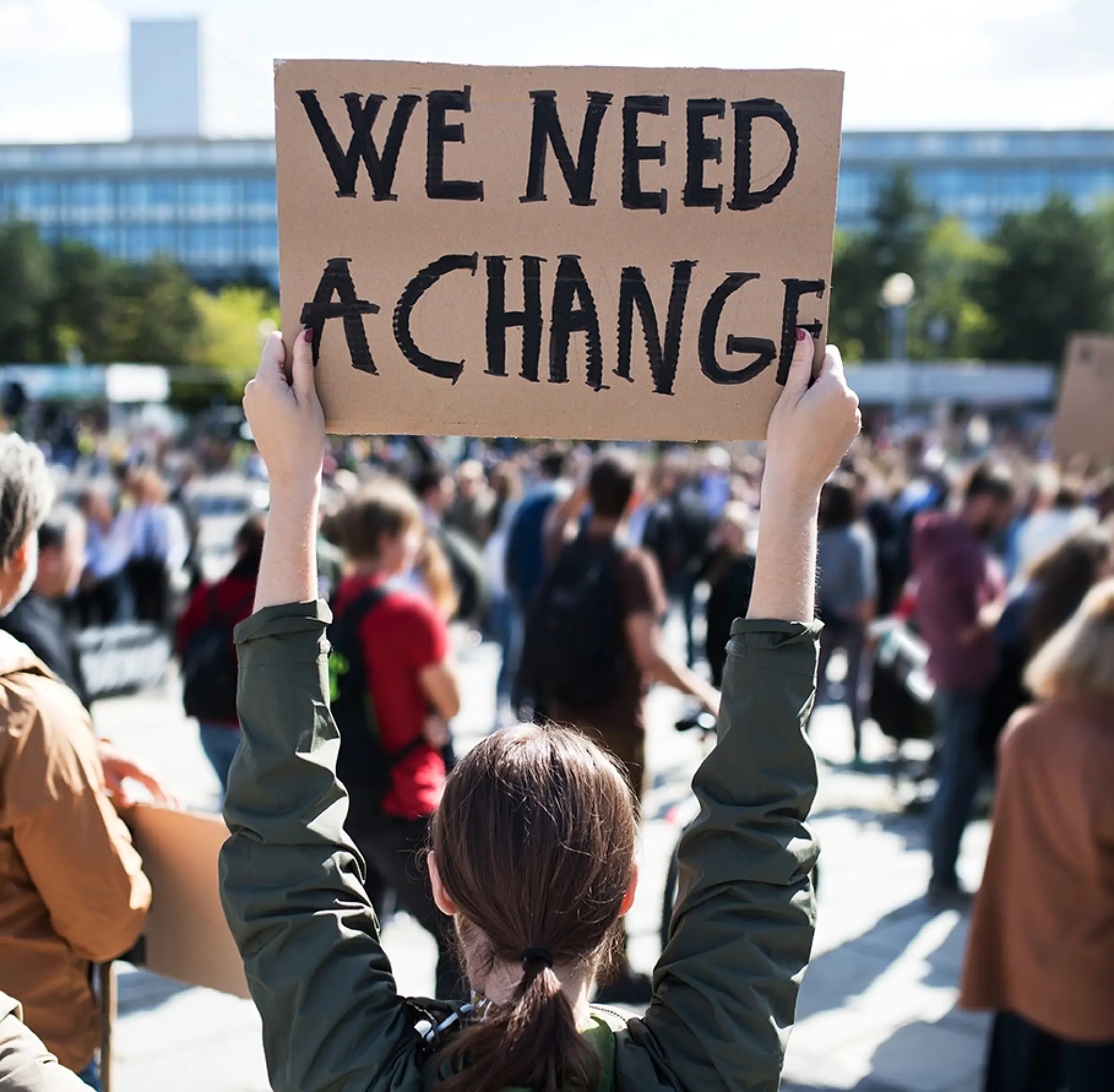 A person in a crowd holds up a cardboard sign reading We need a change. The background shows many people gathered in an outdoor area with trees and a building visible, along with some using tesa tape for various purposes.