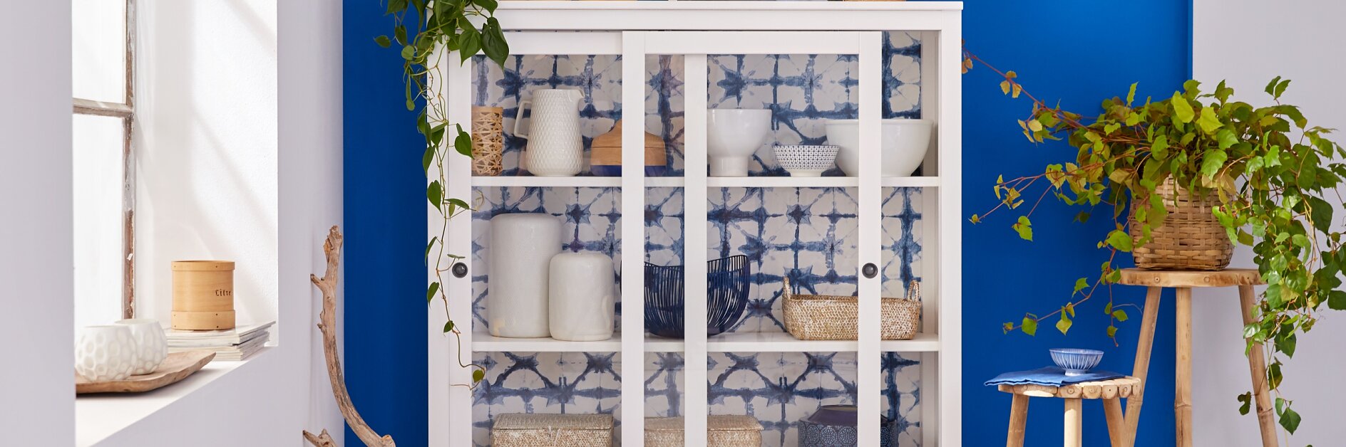 A white cabinet with glass doors stands against a bright blue wall. Inside, various white and wicker items are displayed. A wooden stool with a plant and bowl is beside the cabinet. A leafy branch decorates the area next to a large window.