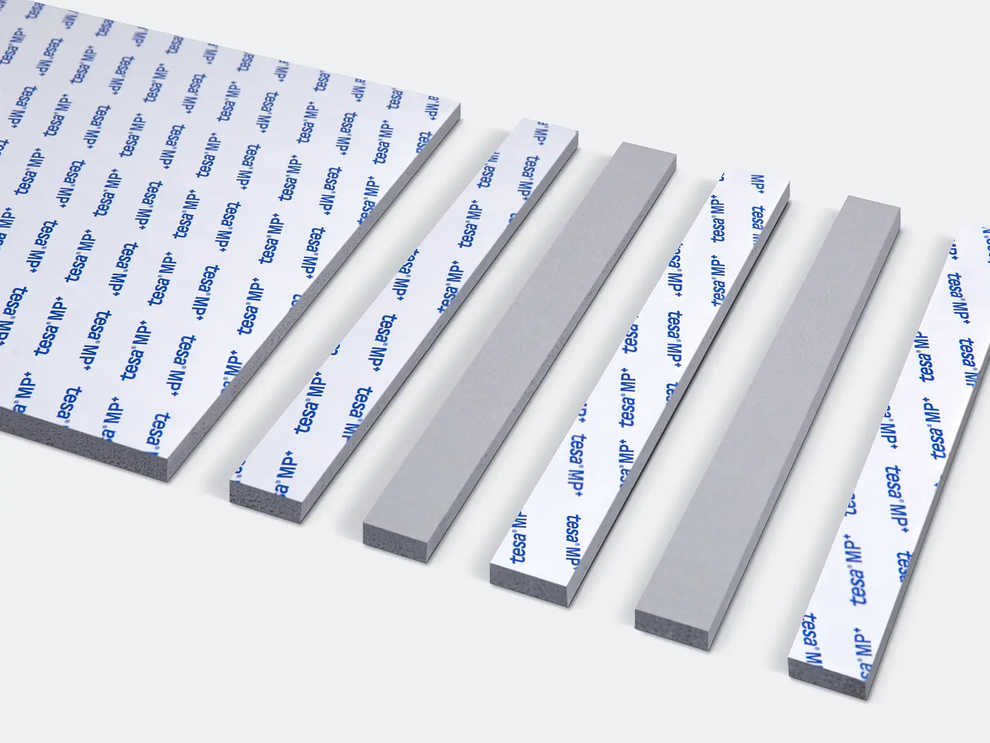Several rectangular foam adhesive strips are laid out on a light gray surface. Each strip has a protective layer with printed text on top, showing varying widths and lengths, arranged parallel to each other.