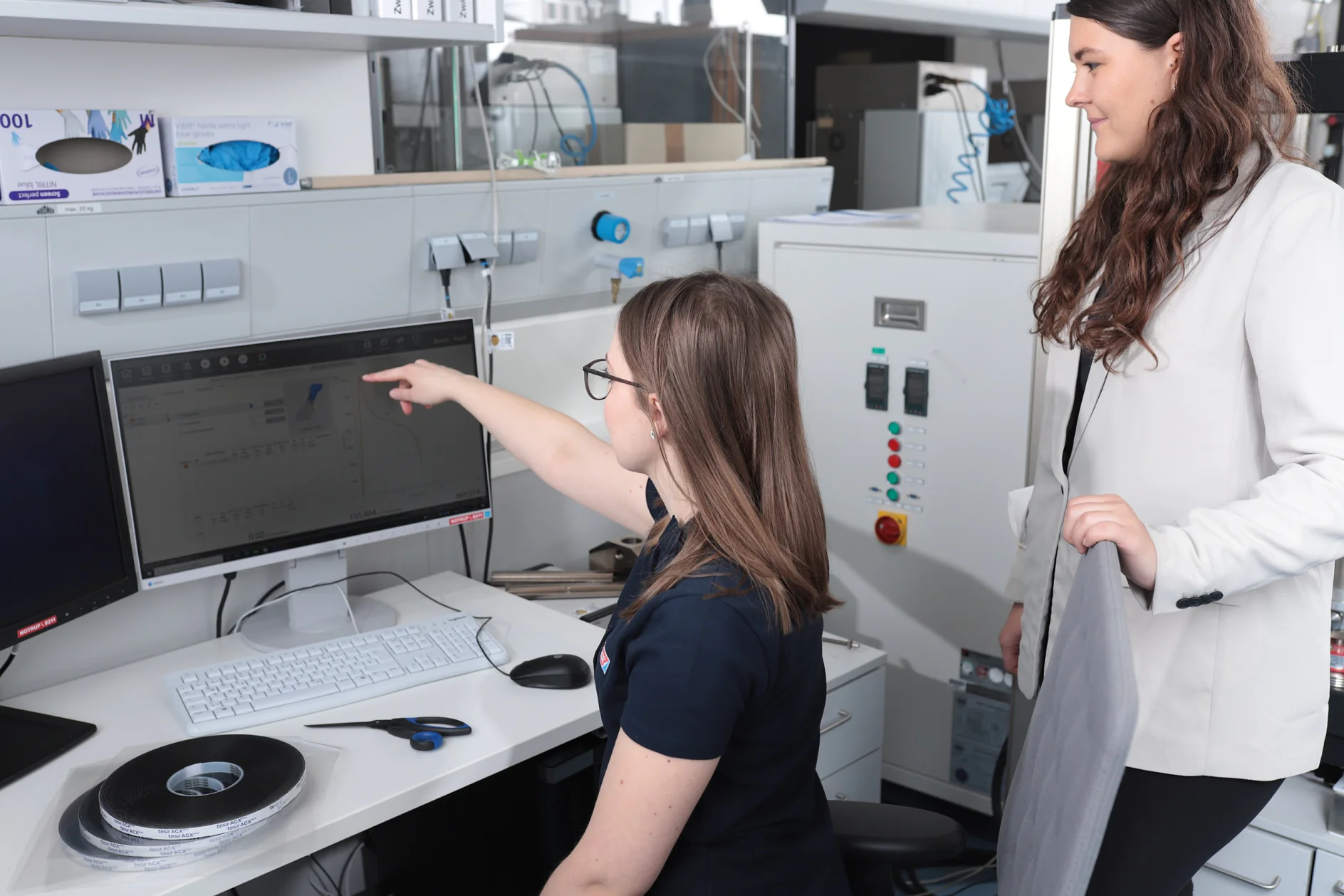 Two women in an office setting. One seated at a desk with a computer, pointing at the monitor. The other woman stands beside her, holding some materials. Office equipment and tesa tape are visible in the background. (This text has been generated by AI)