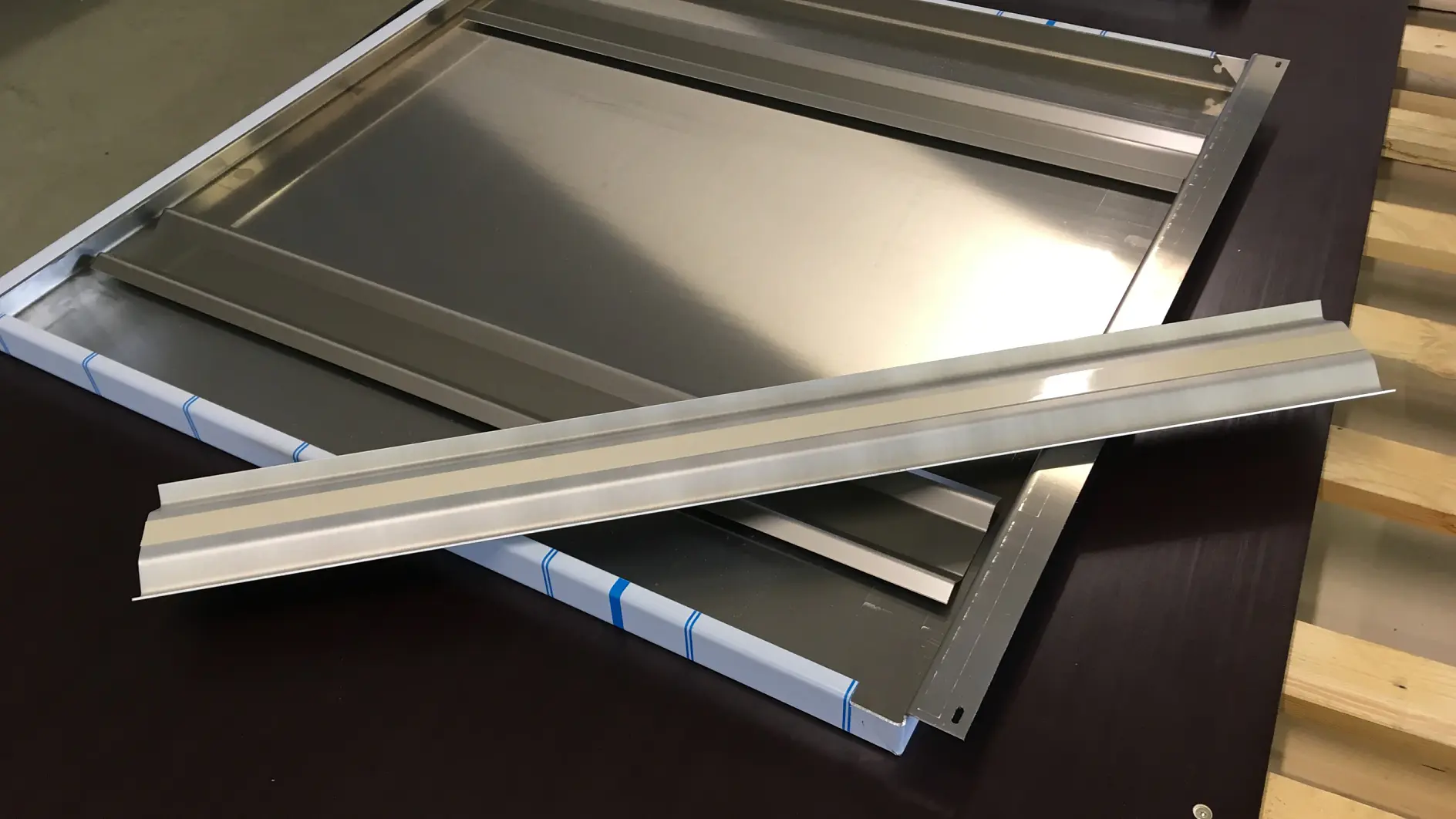 A stack of metallic panels and a separate metal strip lying on a flat surface. The panels appear reflective and are partially wrapped at the edges with tesa tape. The metal strip rests diagonally across the panels. The background includes wooden elements. (This text has been generated by AI)